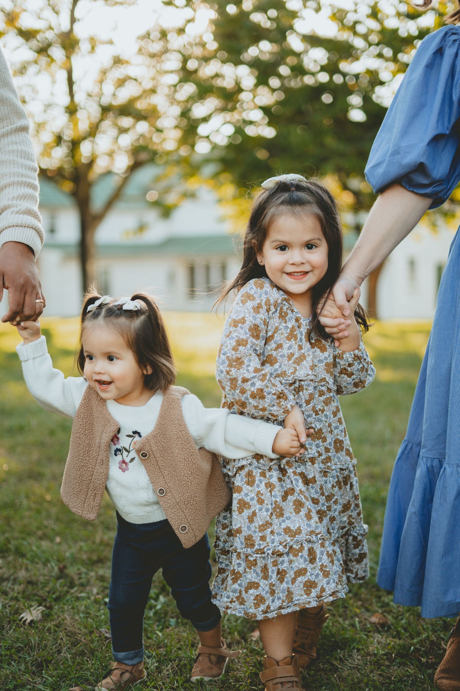 Two young sisters holding parents hands while walking in a sunny park.