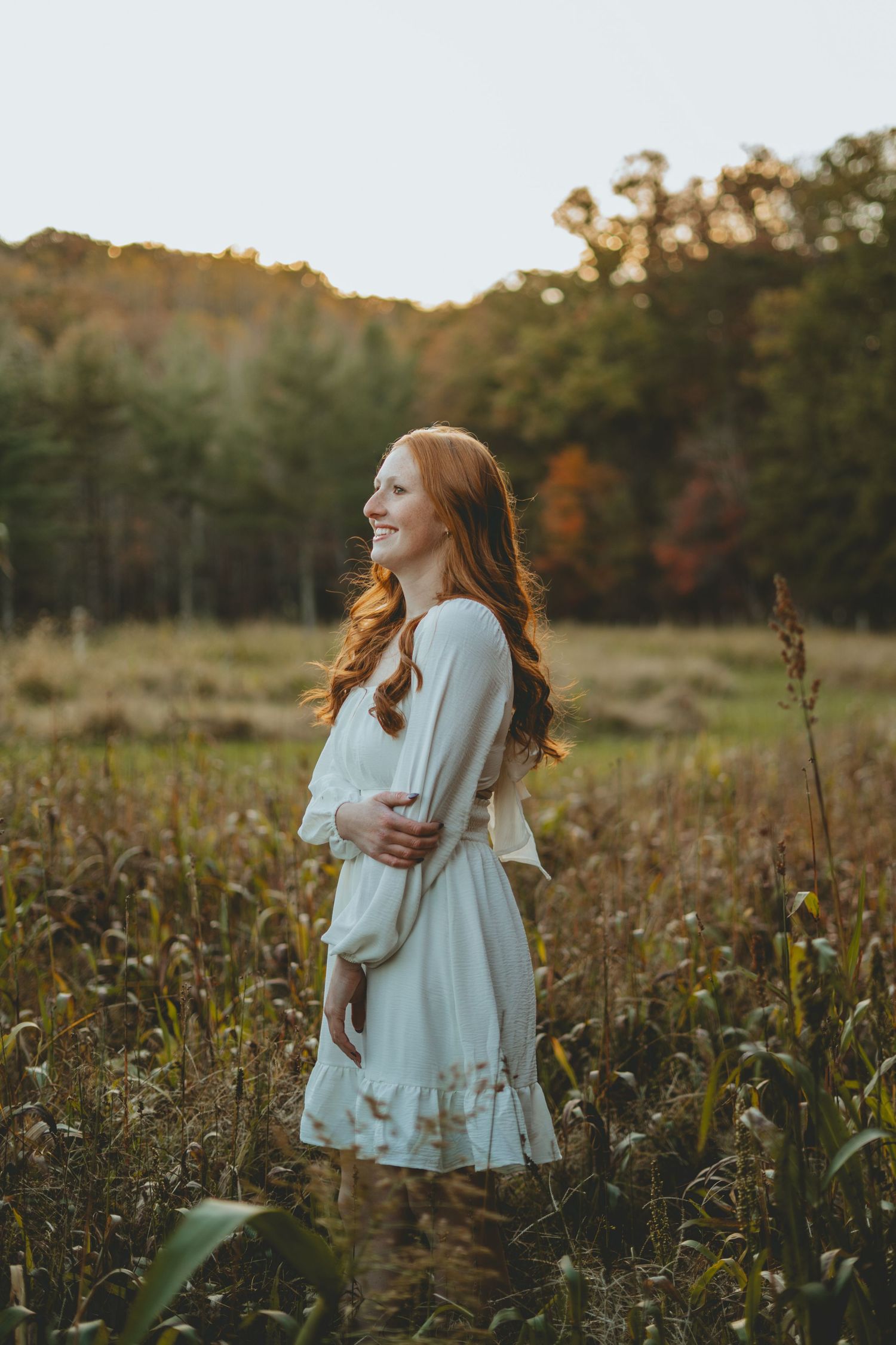 Redhead girl in white dress standing in a sunlit autumn meadow with fall foliage.