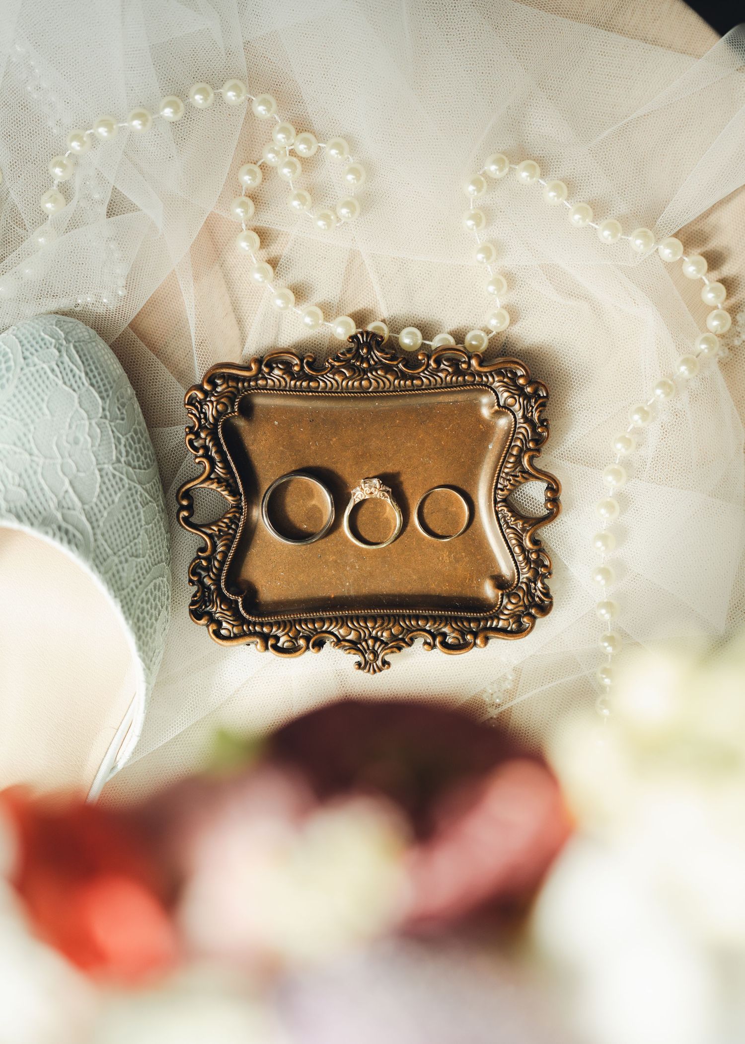 Ornate gold vintage ring dish holding three wedding bands on white lace veil with pearl necklace at wedding.