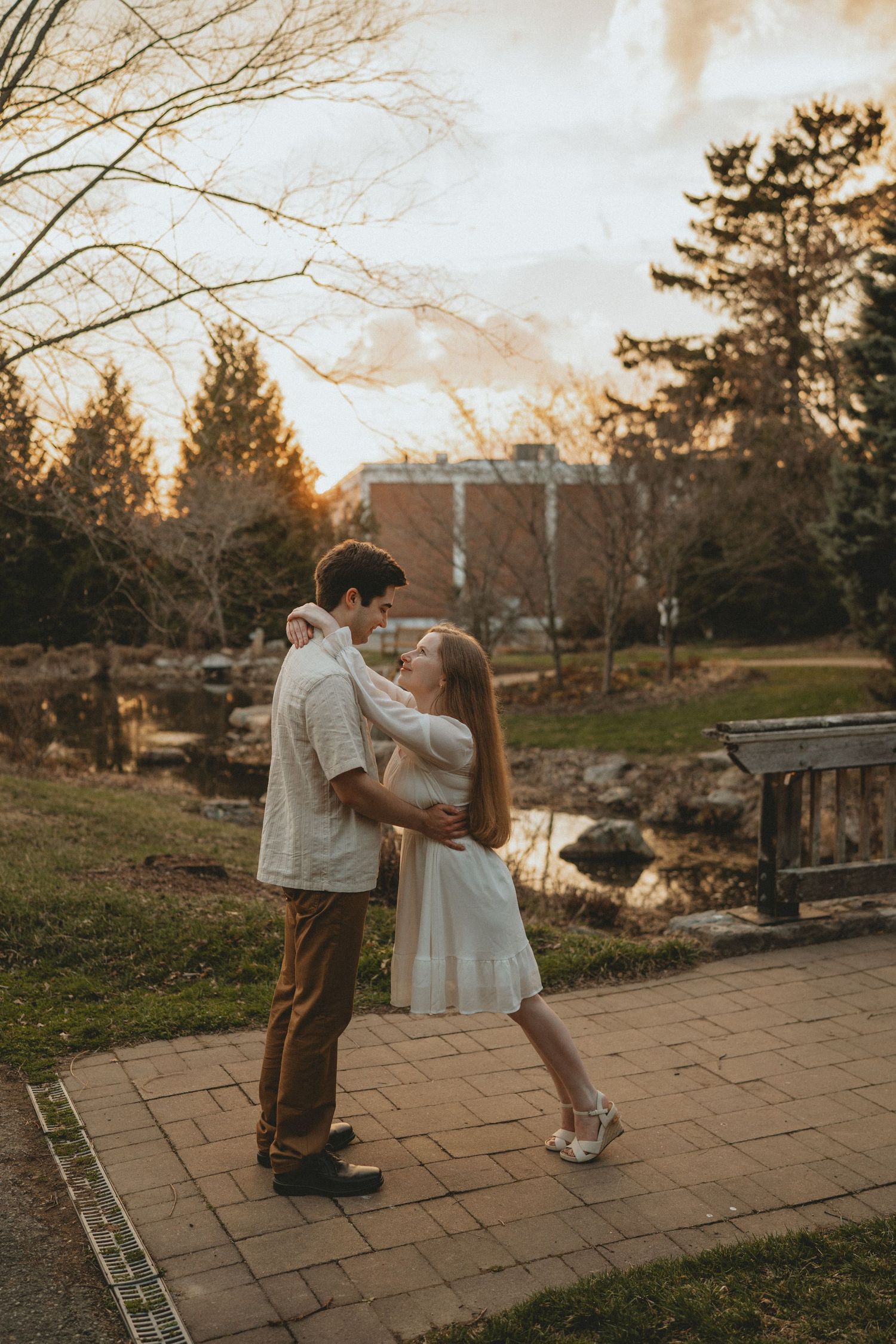 A couple dances together on a garden path at sunset with a pond and trees in the background.