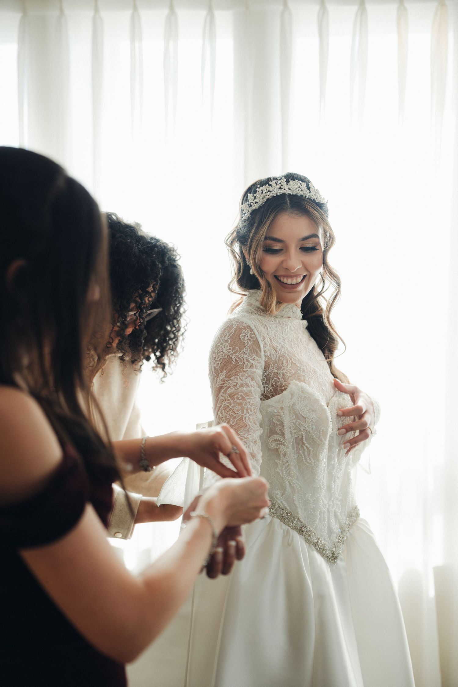 Smiling bride in lace wedding gown and crystal tiara being helped by bridesmaids near bright window.