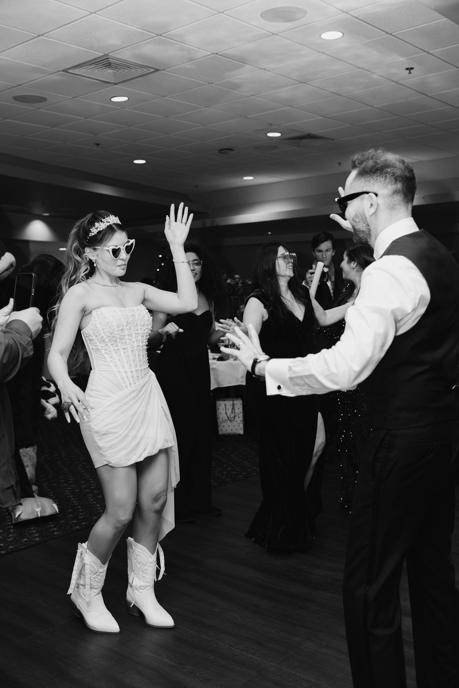 Black and white photo of bride in white mini dress and cowboy boots dancing with groom at wedding reception.
