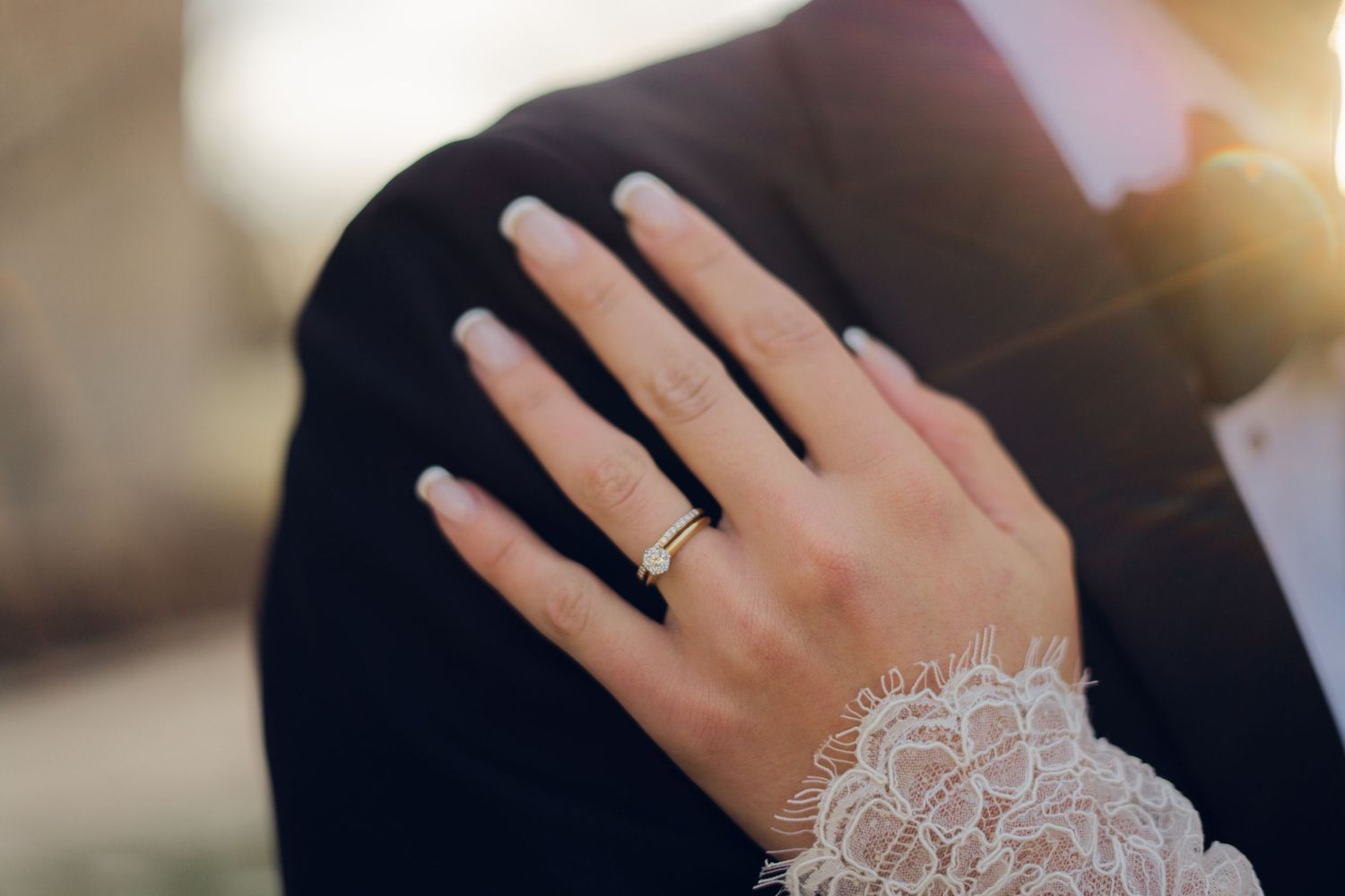 Close-up of bride's hand with gold wedding band resting on groom's black tuxedo jacket at golden hour.