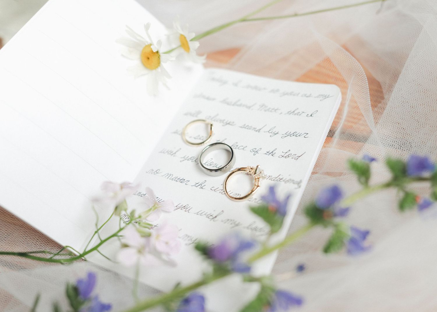 Three wedding rings arranged on handwritten vows surrounded by delicate wildflowers and soft tulle fabric.