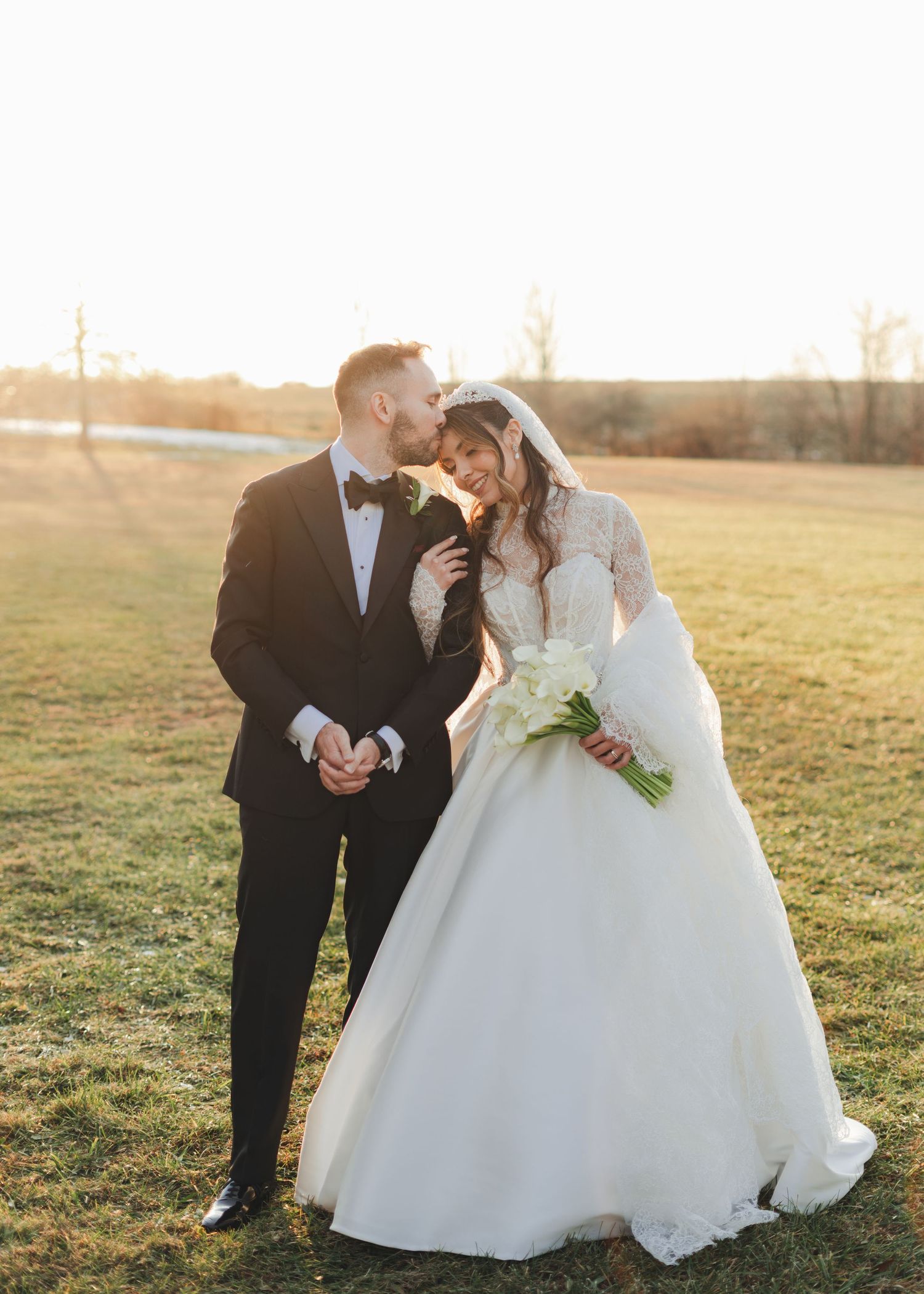 Romantic couple shares tender moment at golden sunset, groom in tuxedo kisses bride in lace ball gown.