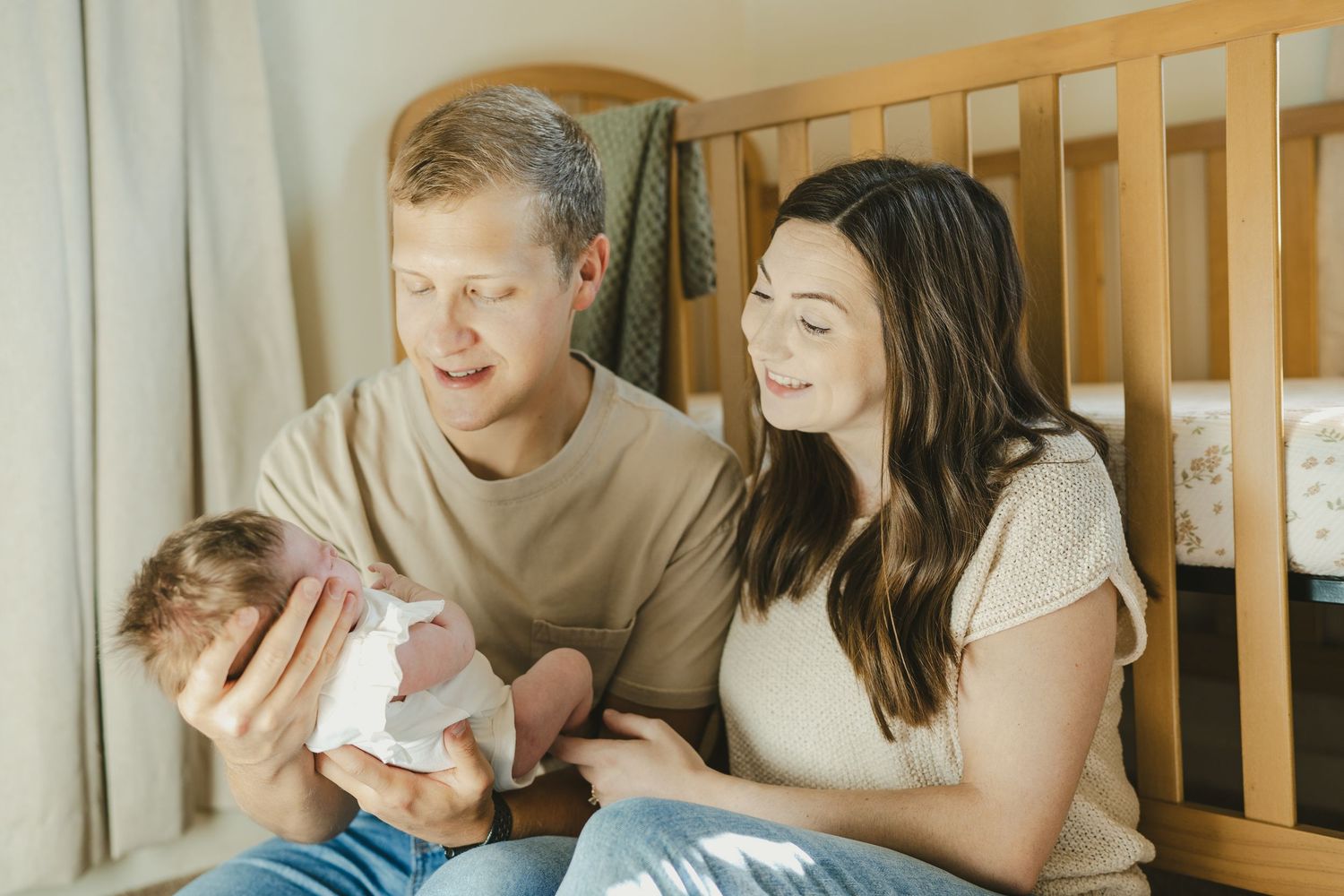 Smiling parents sitting by wooden crib holding their newborn baby.