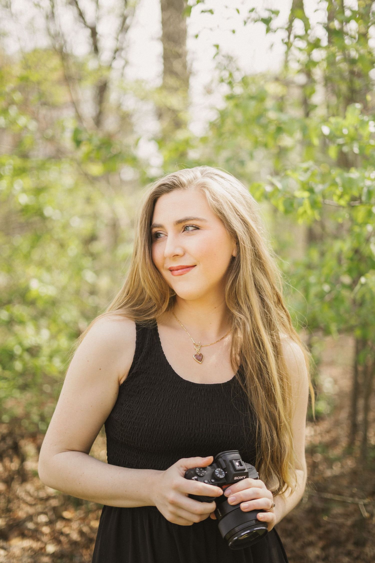 Young blonde woman holding camera in sunlit forest wearing black dress.