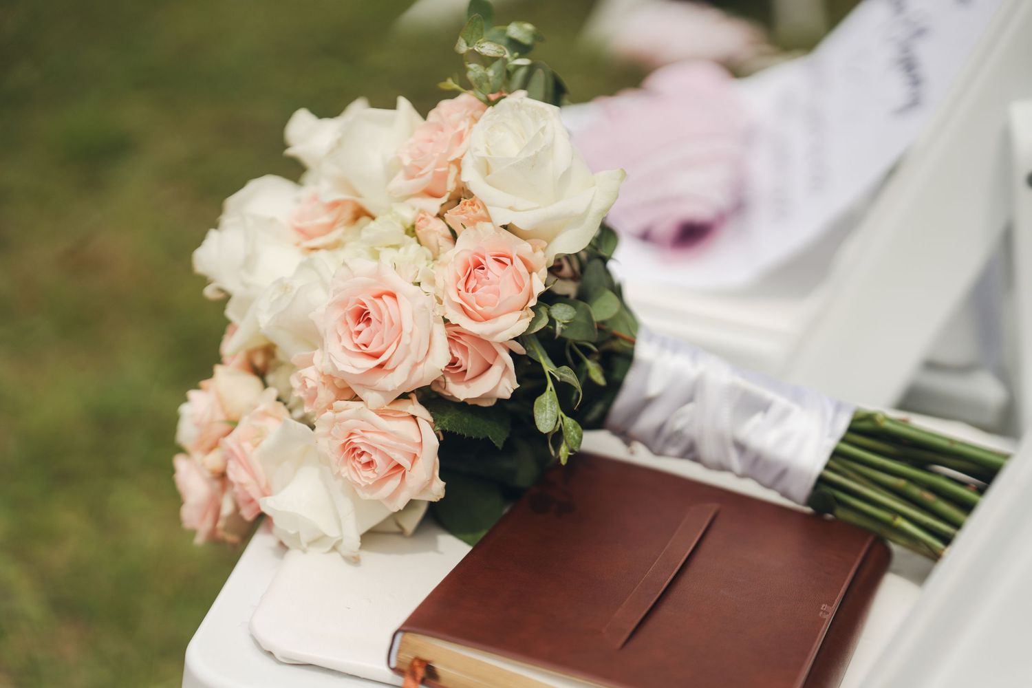 Elegant wedding bouquet of blush and white roses resting on a brown leather book outdoors.