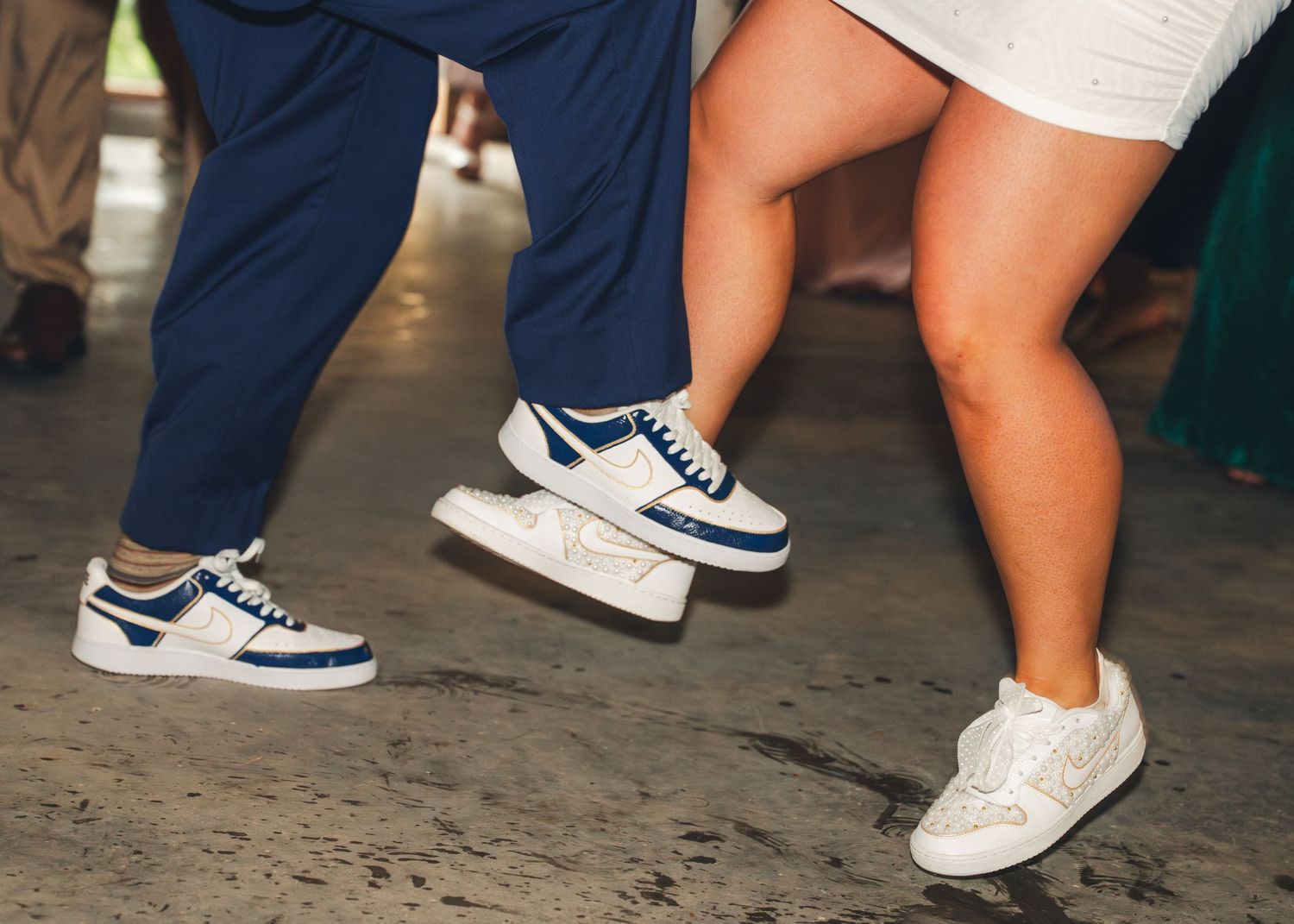 Two people dancing in white and blue sneakers on a concrete floor at a casual event.