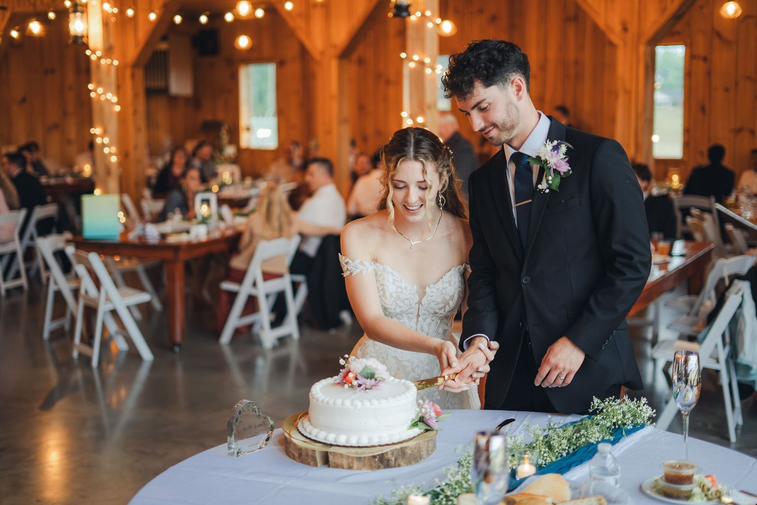 Newlyweds cut their white wedding cake together in a warmly lit rustic barn reception hall filled with guests.