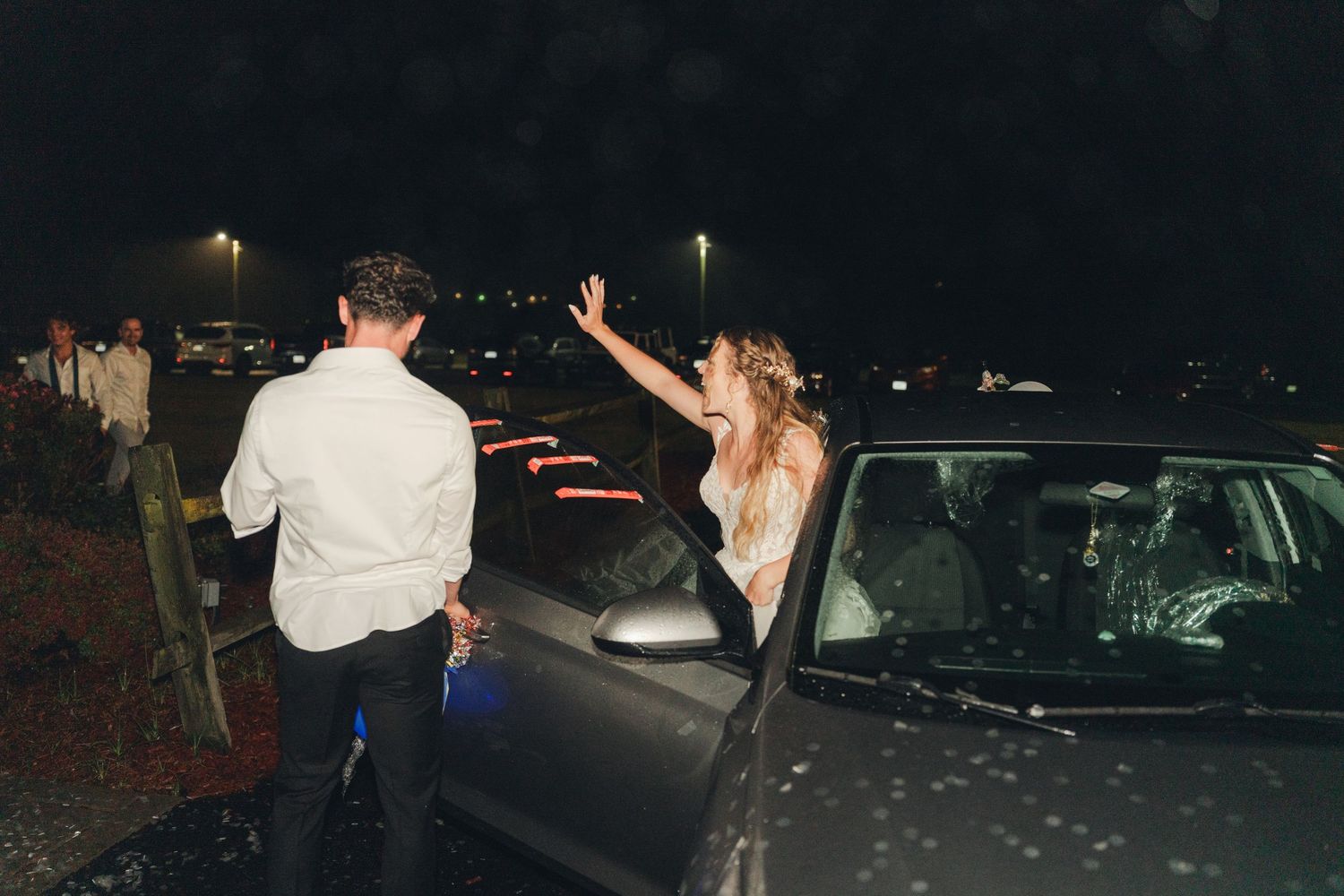 Bride and groom celebrate beside a decorated car at night after their wedding reception.
