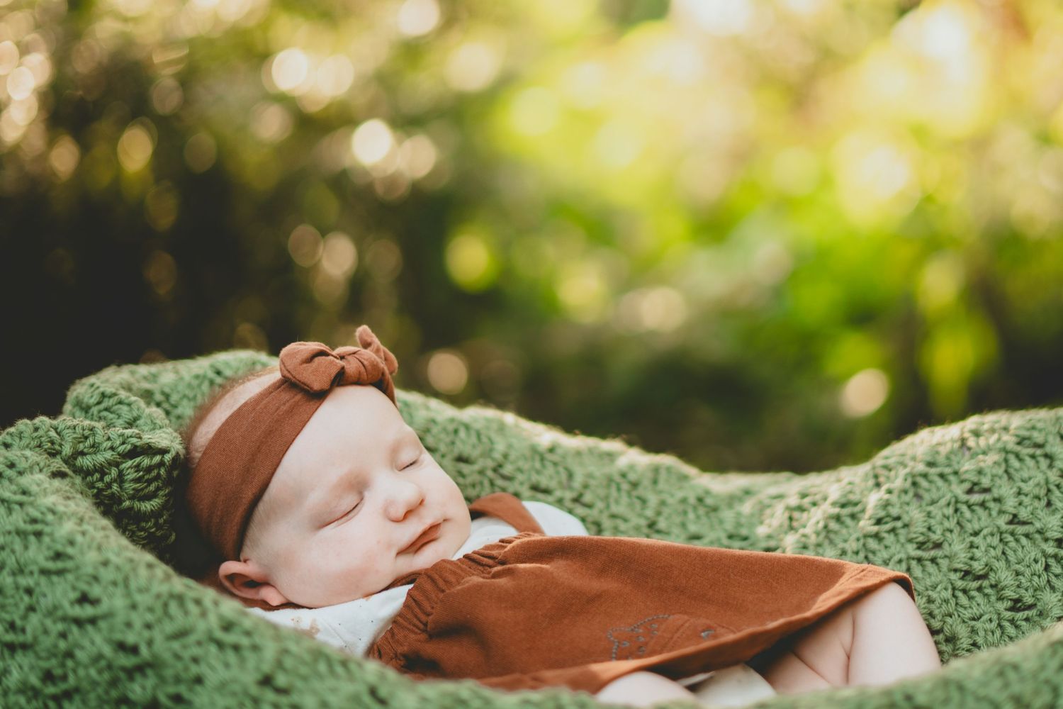 A serene newborn baby sleeps peacefully on a green knit blanket wearing a brown outfit and headband.