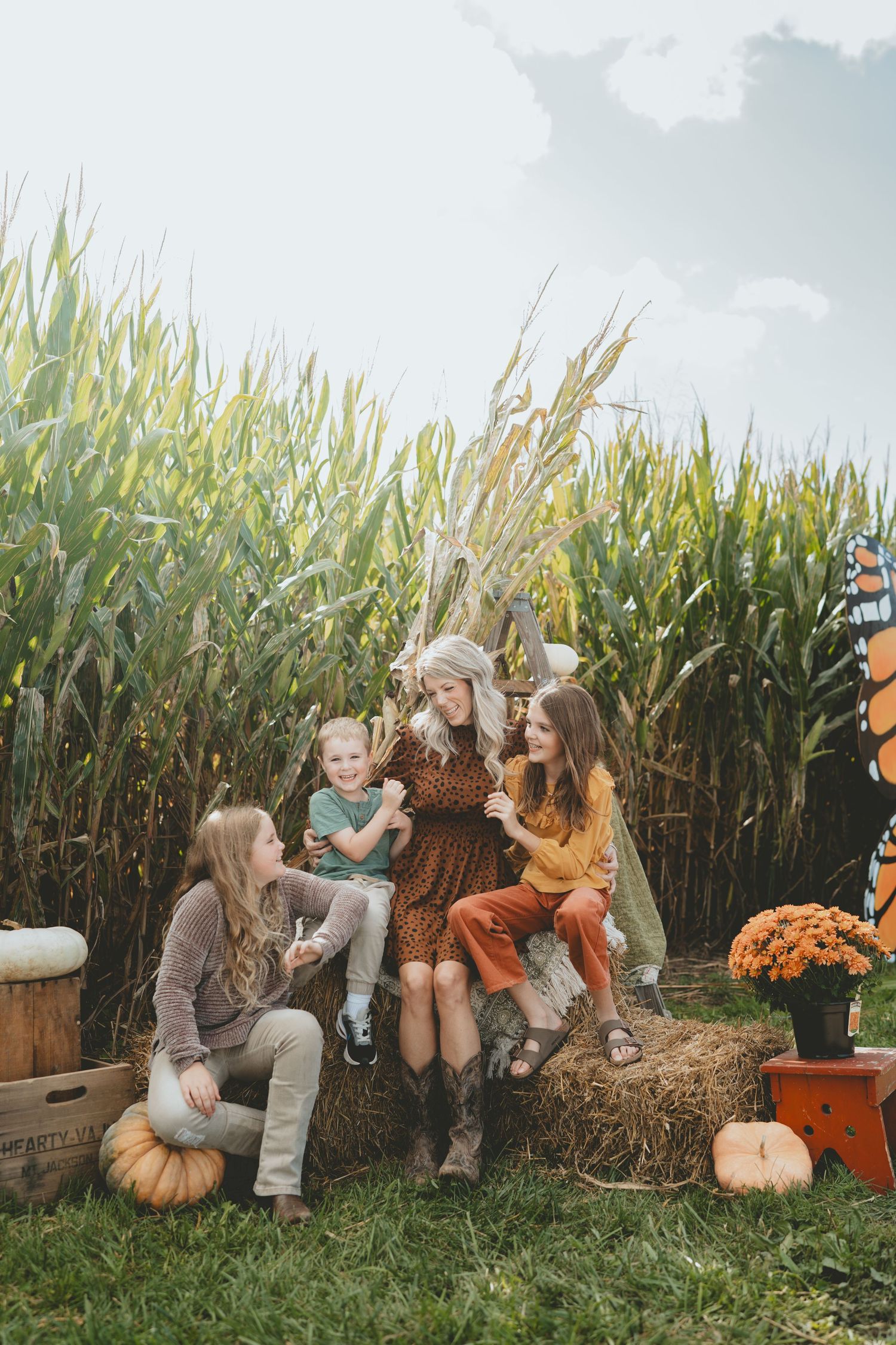 Woman in brown dress with three children on hay bale in front of cornfield.