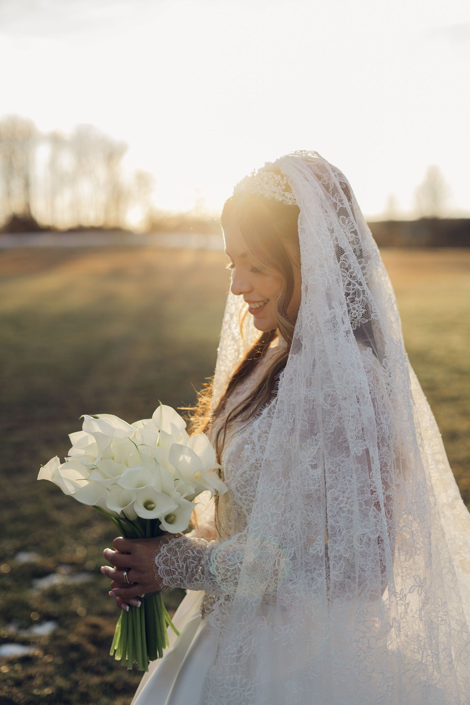 Bride in lace veil holding white calla lily bouquet at golden sunset.