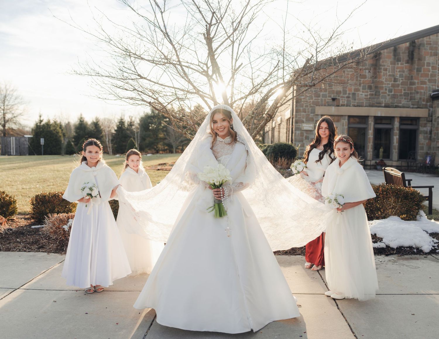 Bride in white gown with veil walking outdoors with flower girls in white dresses in winter.
