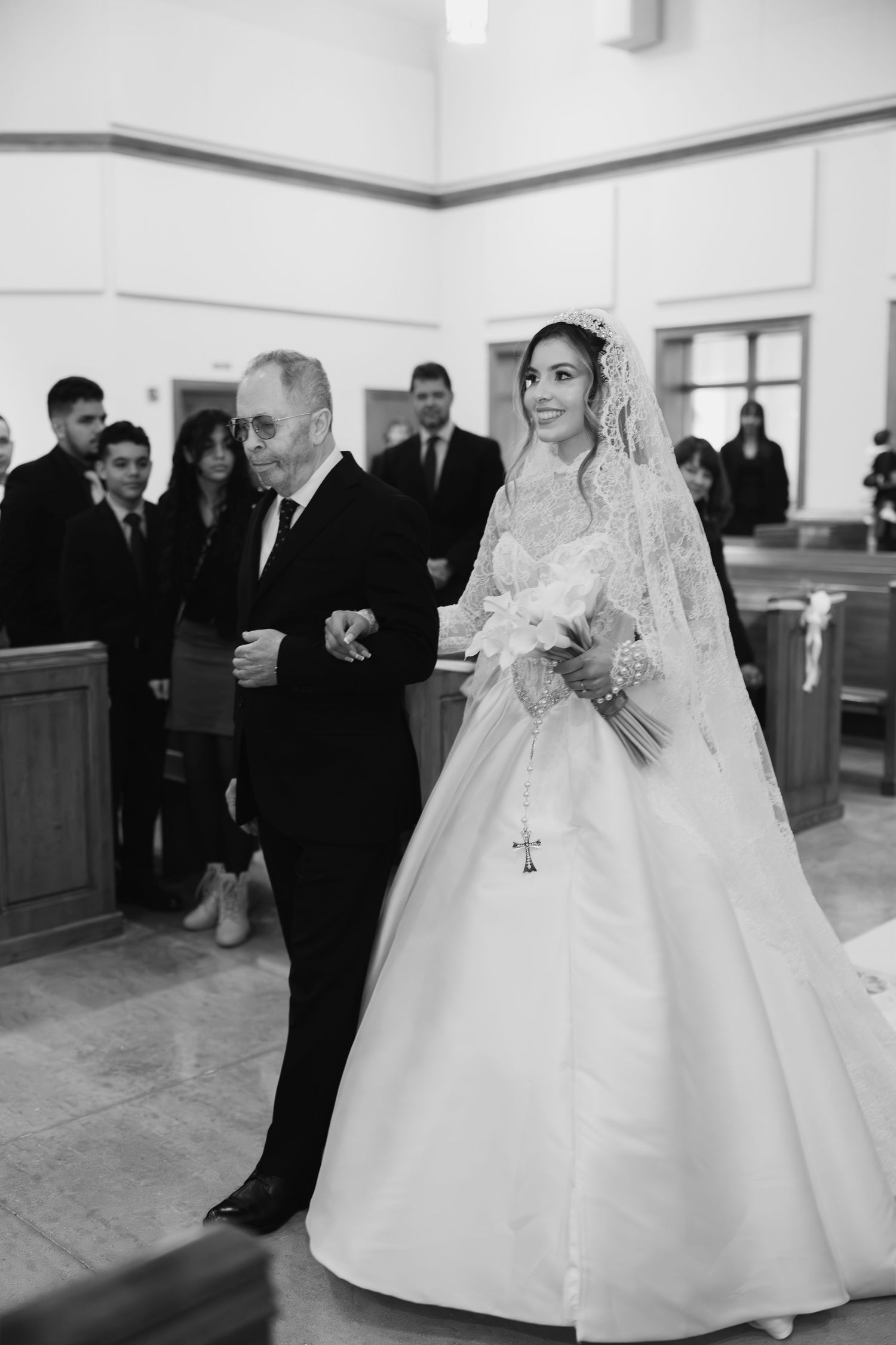 Black and white photo of bride in lace gown and veil walking down church aisle with father.