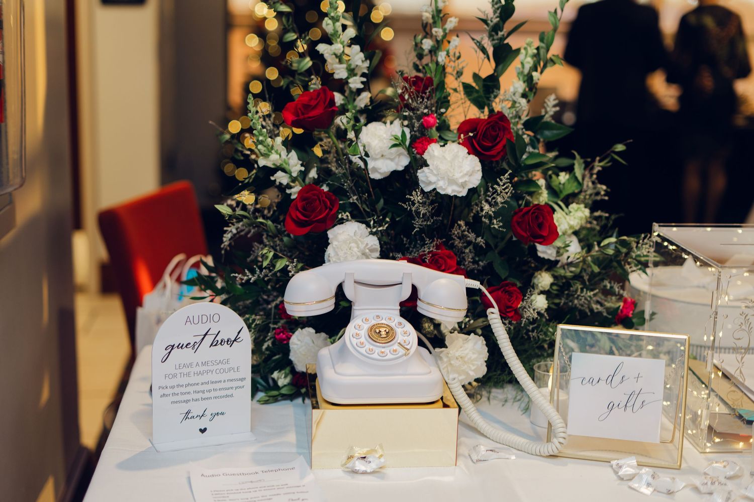 Wedding guest book table with vintage white telephone, red and white floral arrangement, and signage.