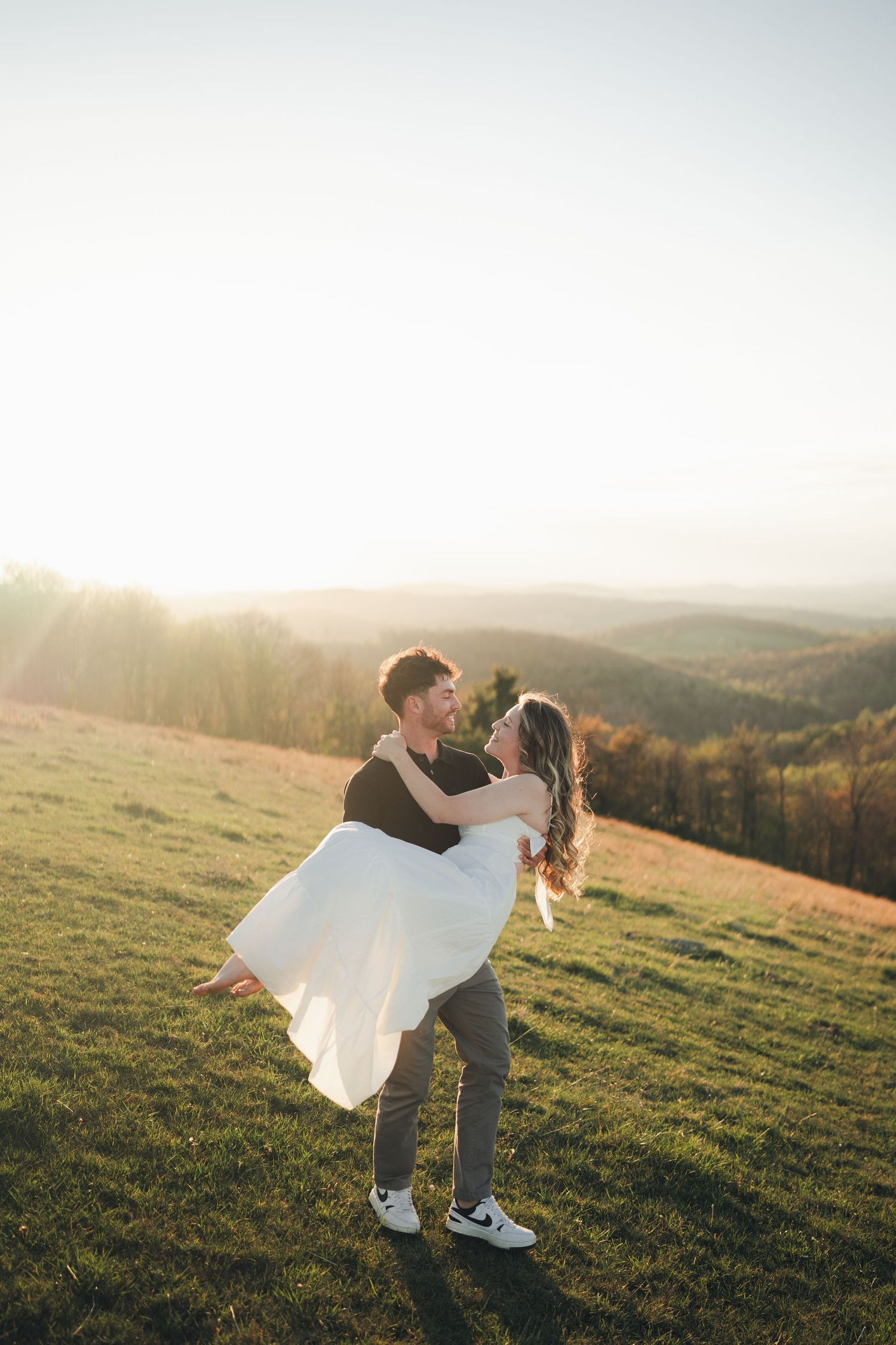 Man lifts smiling woman in white dress at golden sunset over rolling mountain hills during engagement session.