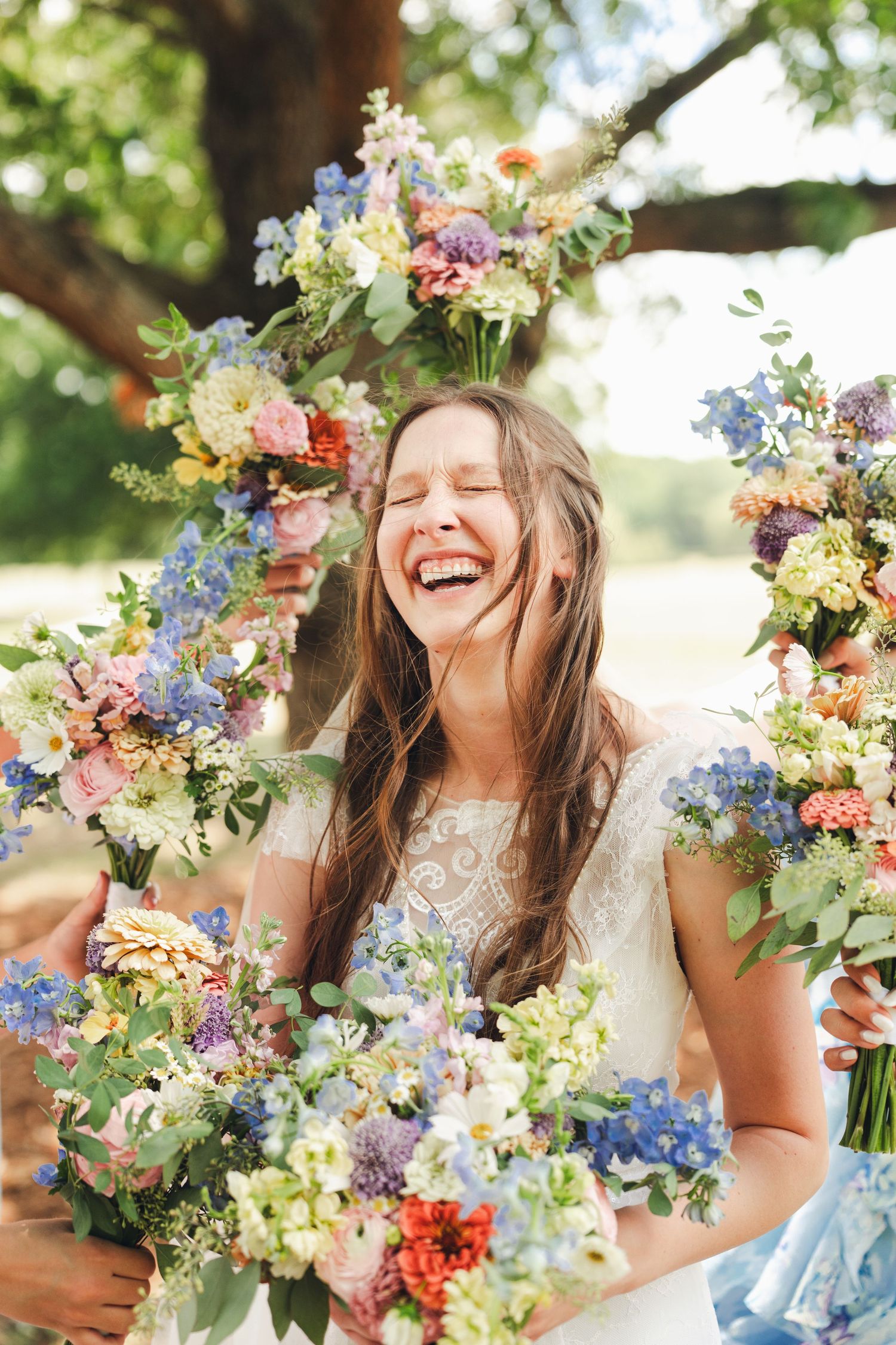Laughing bride in lace wedding dress surrounded by colorful wildflower bouquets outdoors.