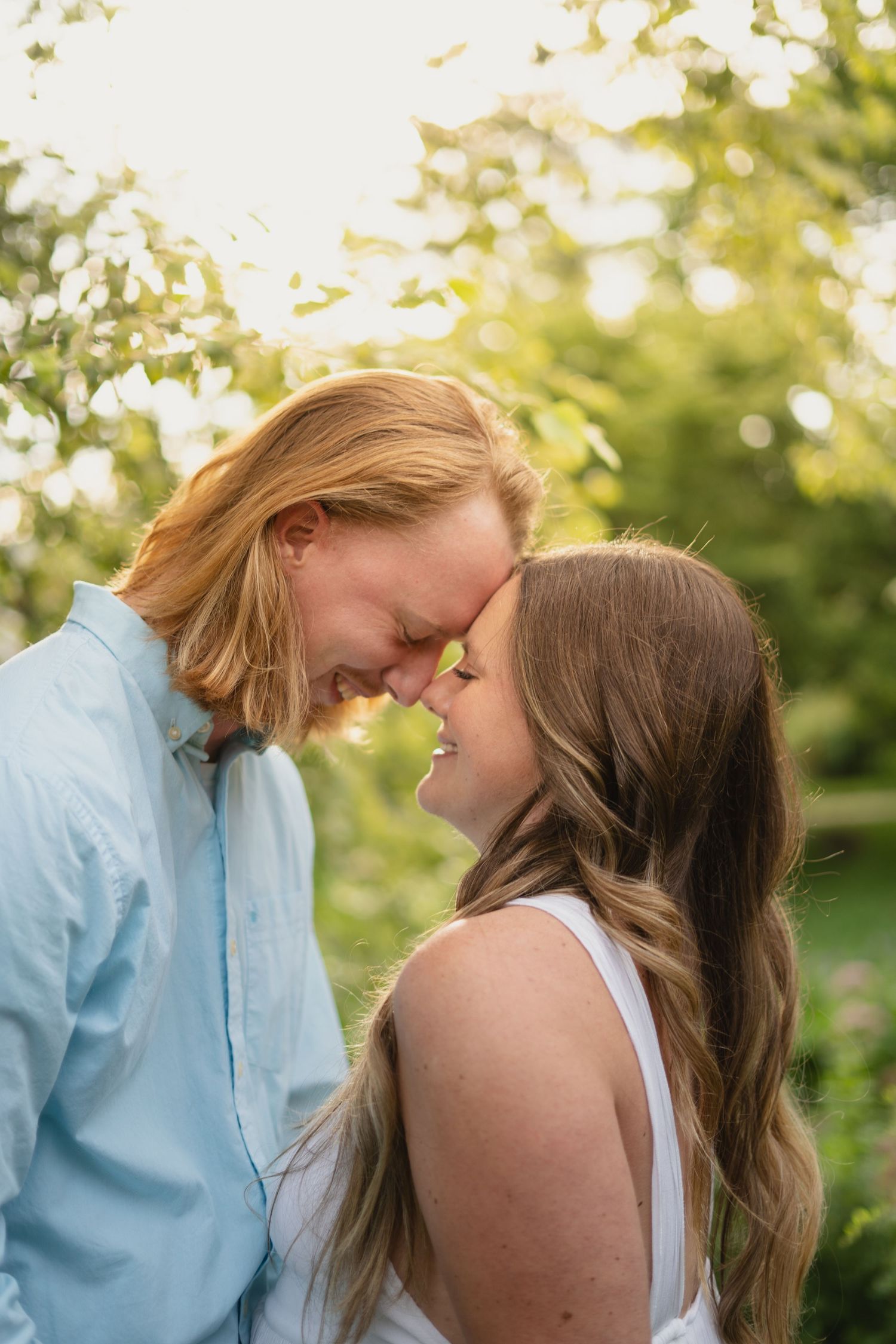 Joyful couple touch foreheads and smile together in golden sunlit green garden background.