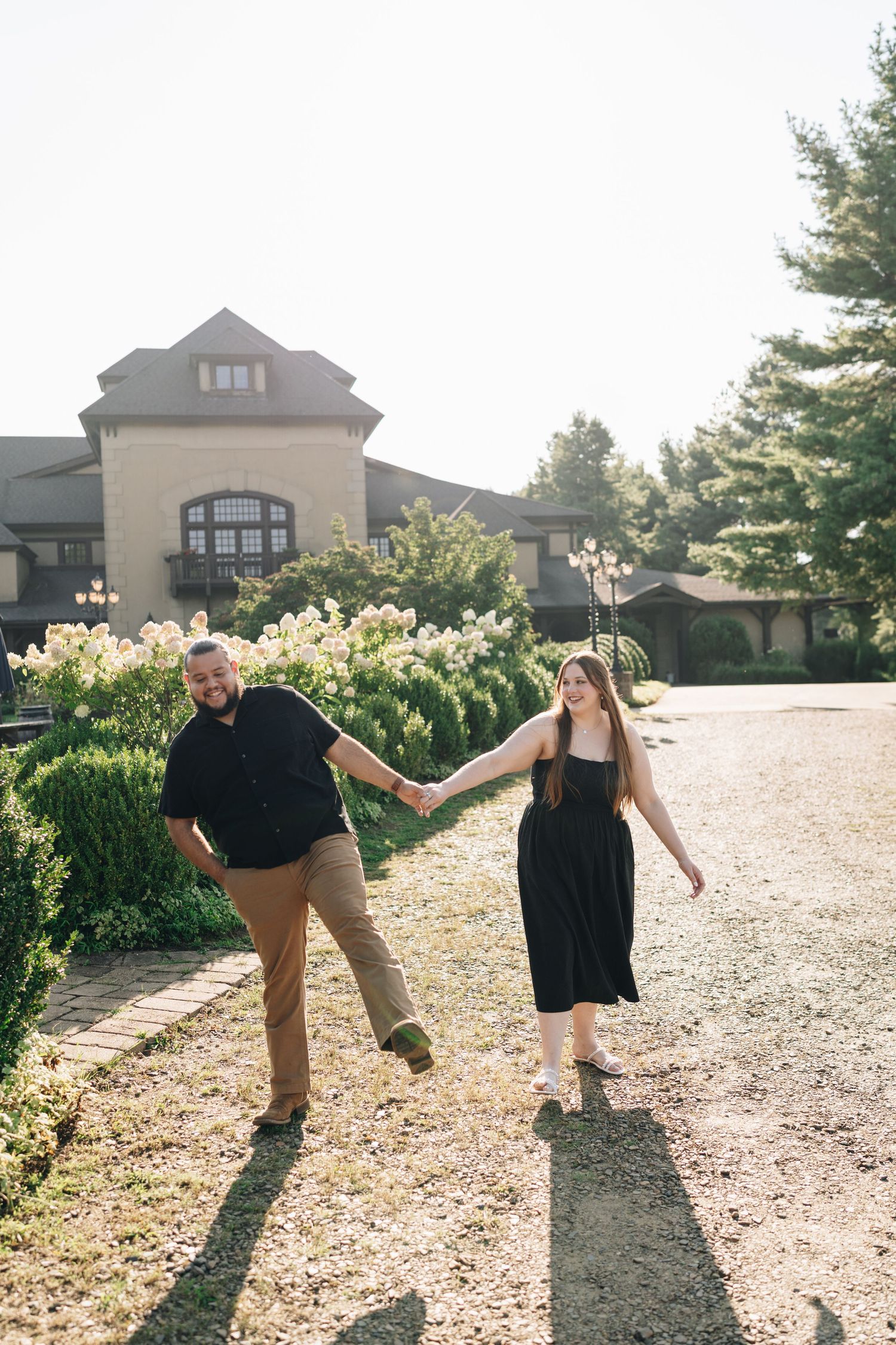Happy couple holding hands and walking along a sunlit garden path near a large estate building.