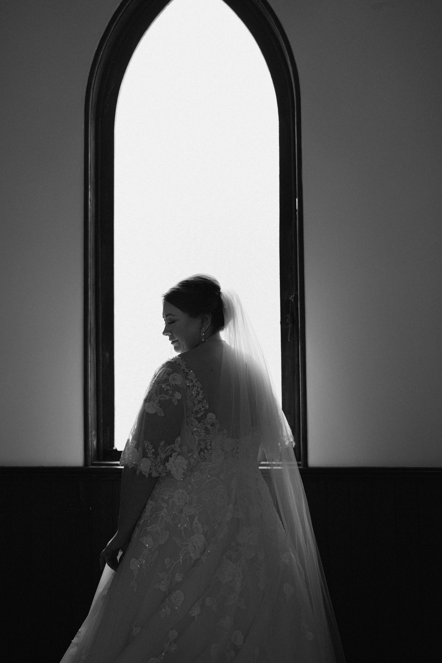 Bride in lace wedding gown and veil silhouetted against arched church window in black and white photo.