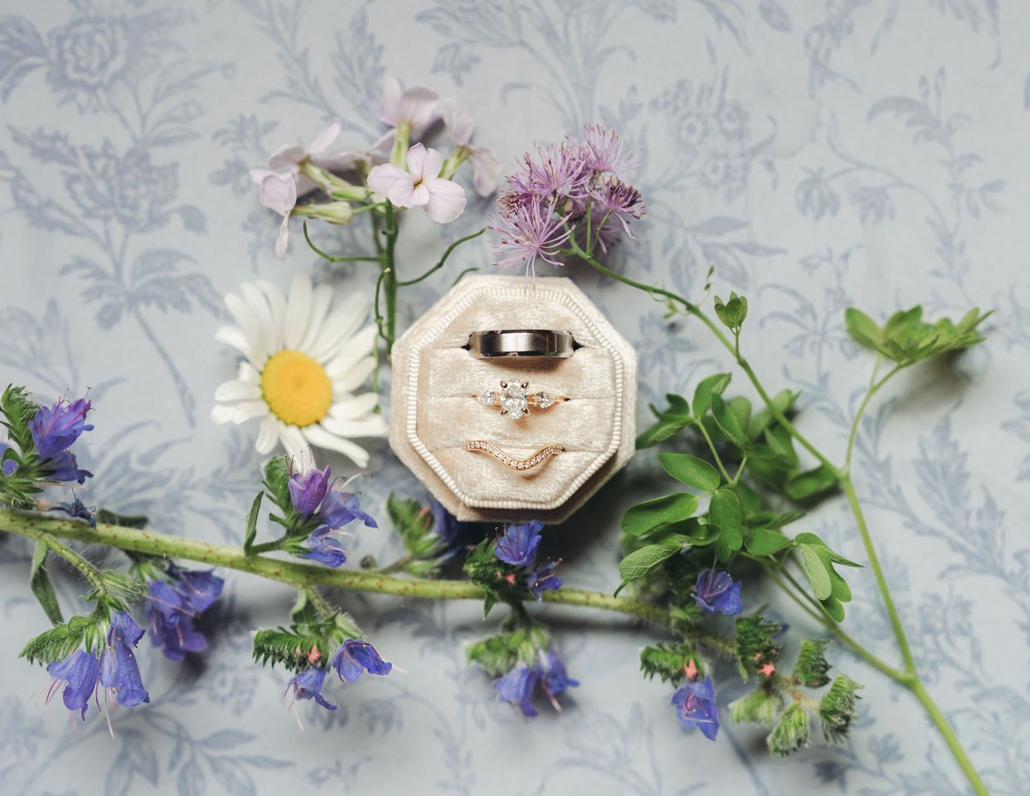 Wedding rings in an octagonal velvet box surrounded by wildflowers including daisies and bluebells on floral fabric.