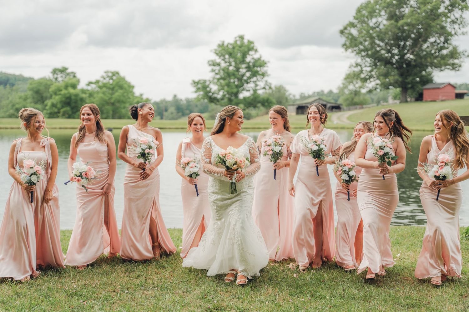 Bride and bridesmaids in blush pink gowns holding bouquets on green lawn near pond at outdoor wedding.