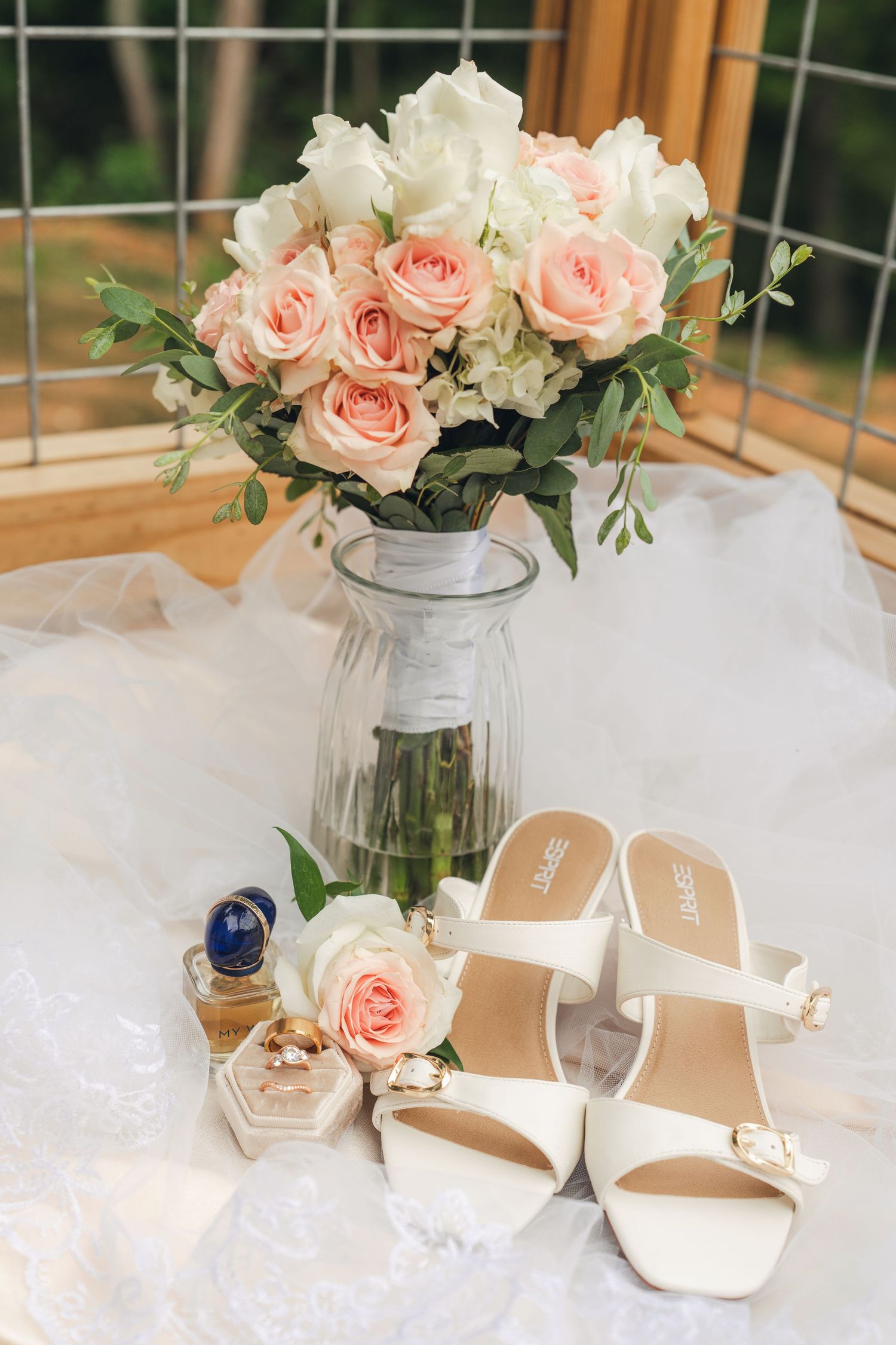 Bridal flat lay featuring white heels, pink roses bouquet in glass vase, gold rings, and perfume on white lace fabric.