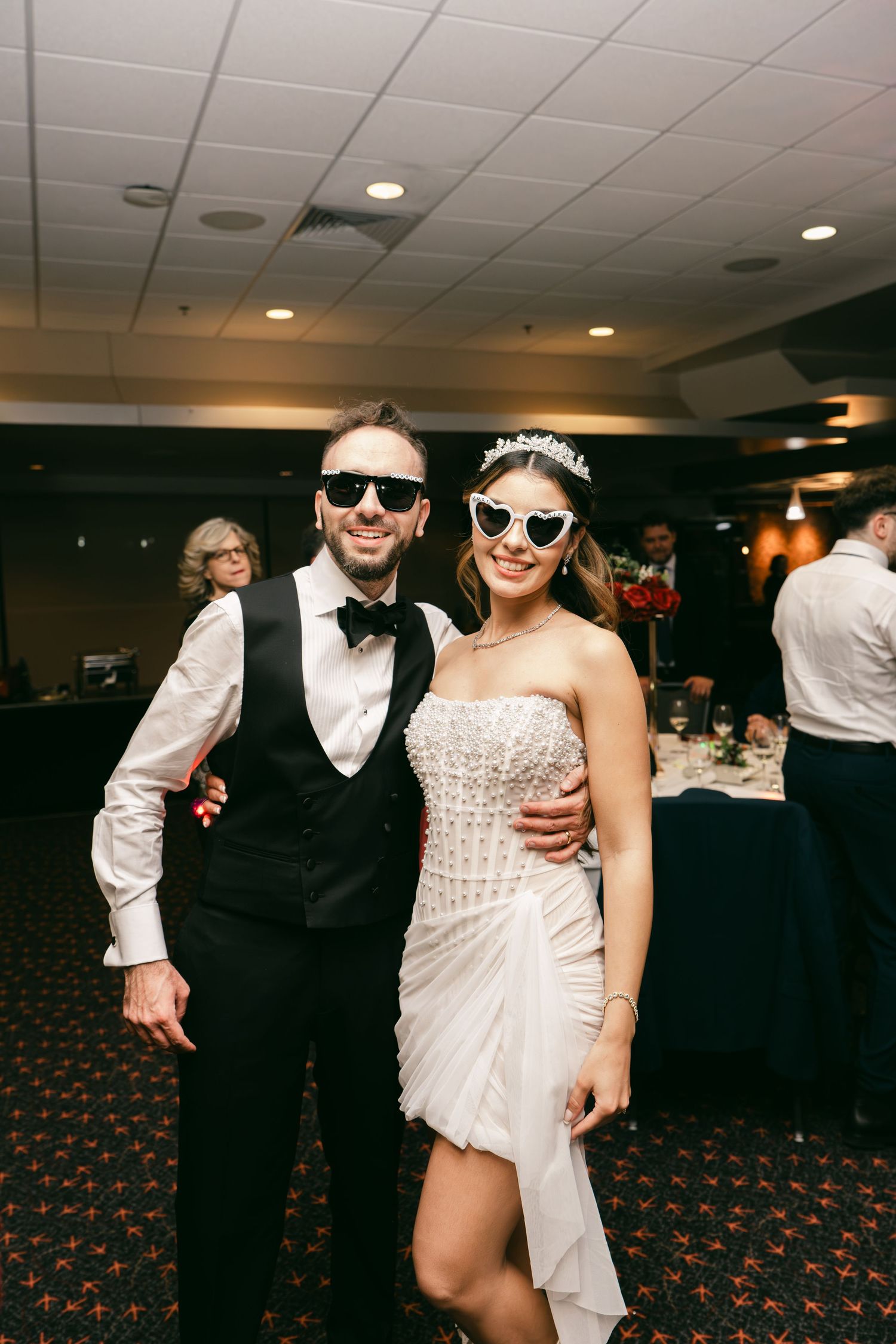 Bride and groom pose playfully at wedding reception wearing matching sunglasses, bride in short white dress with tiara.