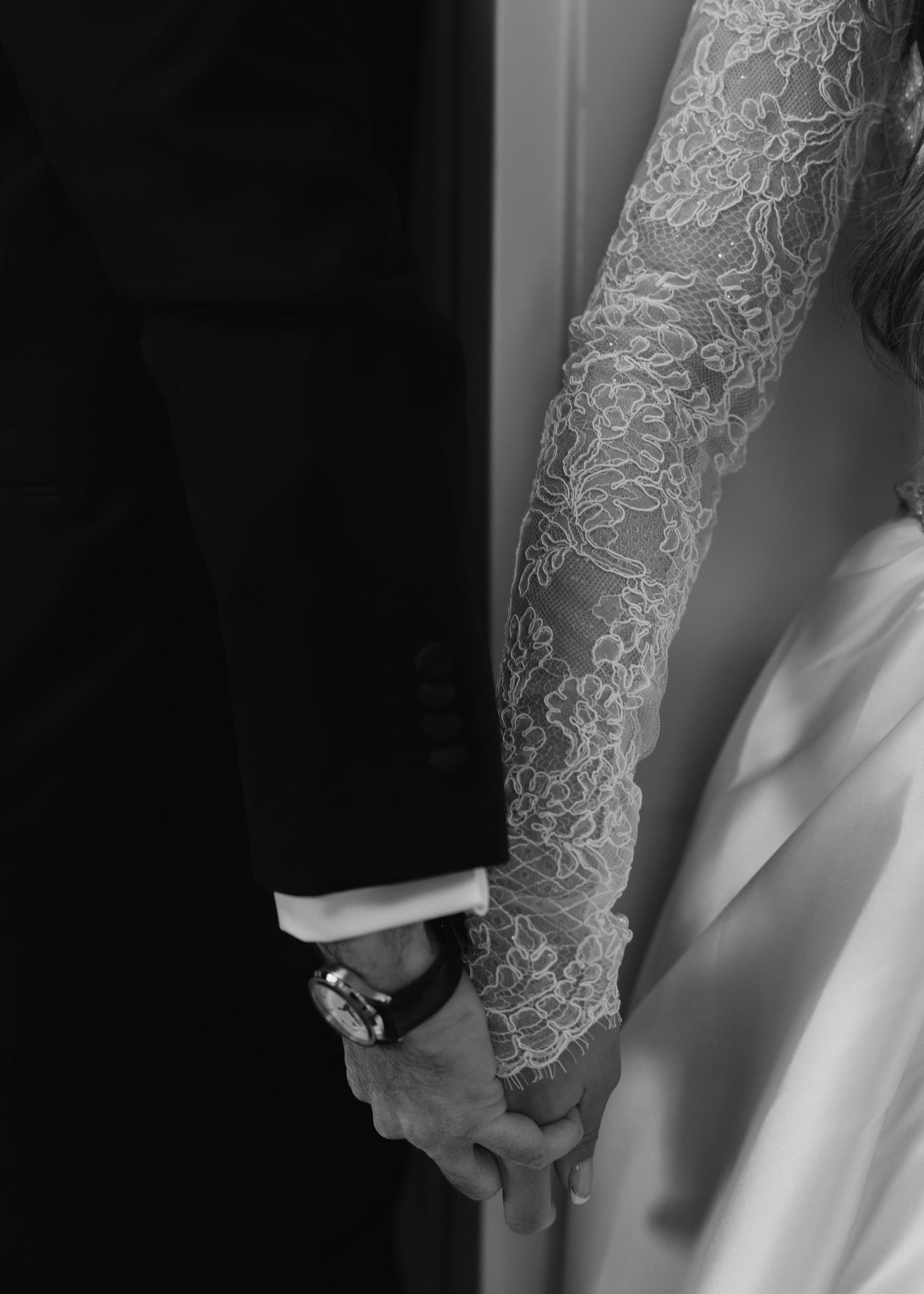 Black and white close-up of bride and groom holding hands, showcasing intricate lace wedding dress sleeve.