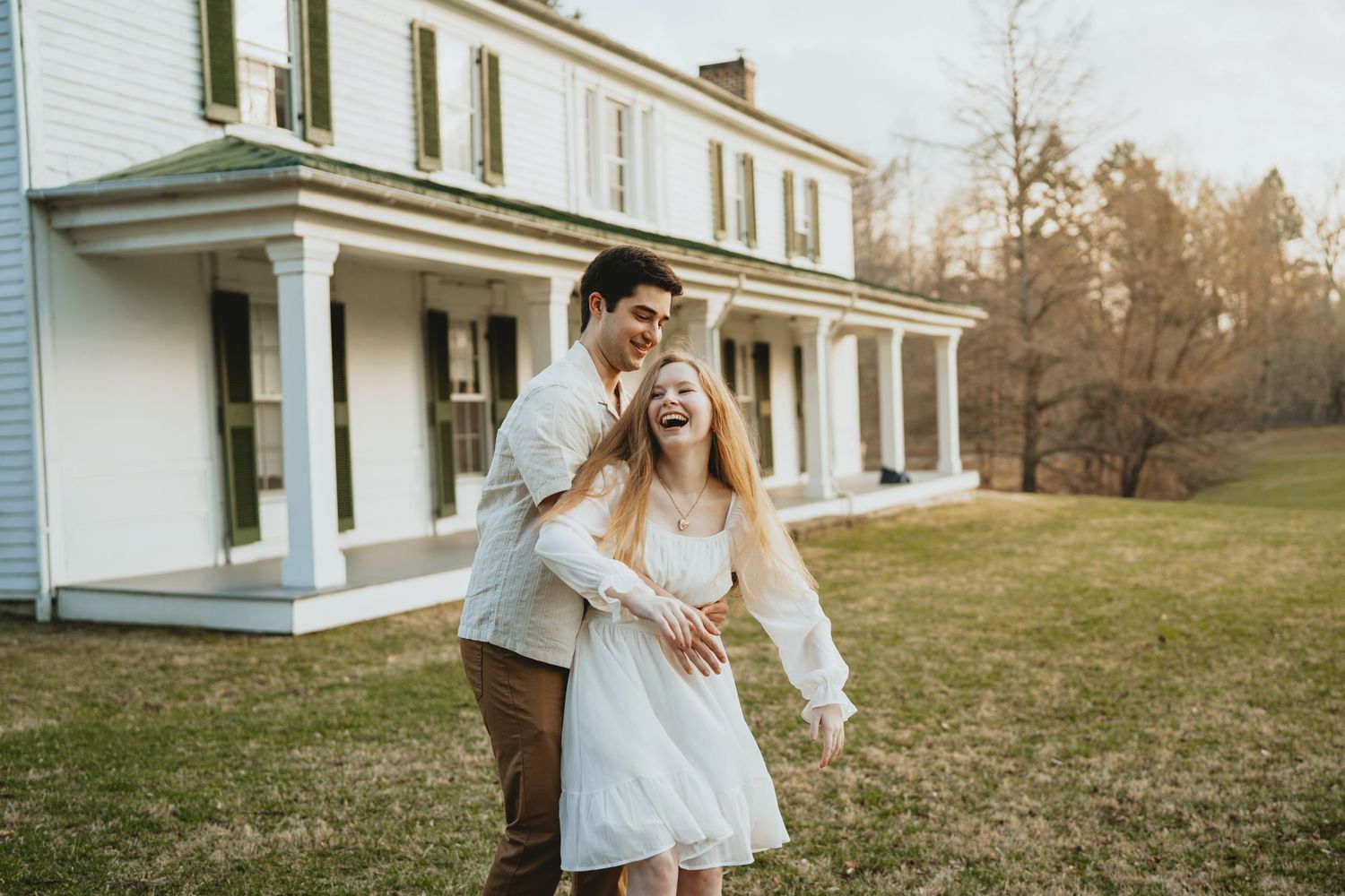 Joyful couple laughing and embracing outside a historic white colonial farmhouse at sunset.
