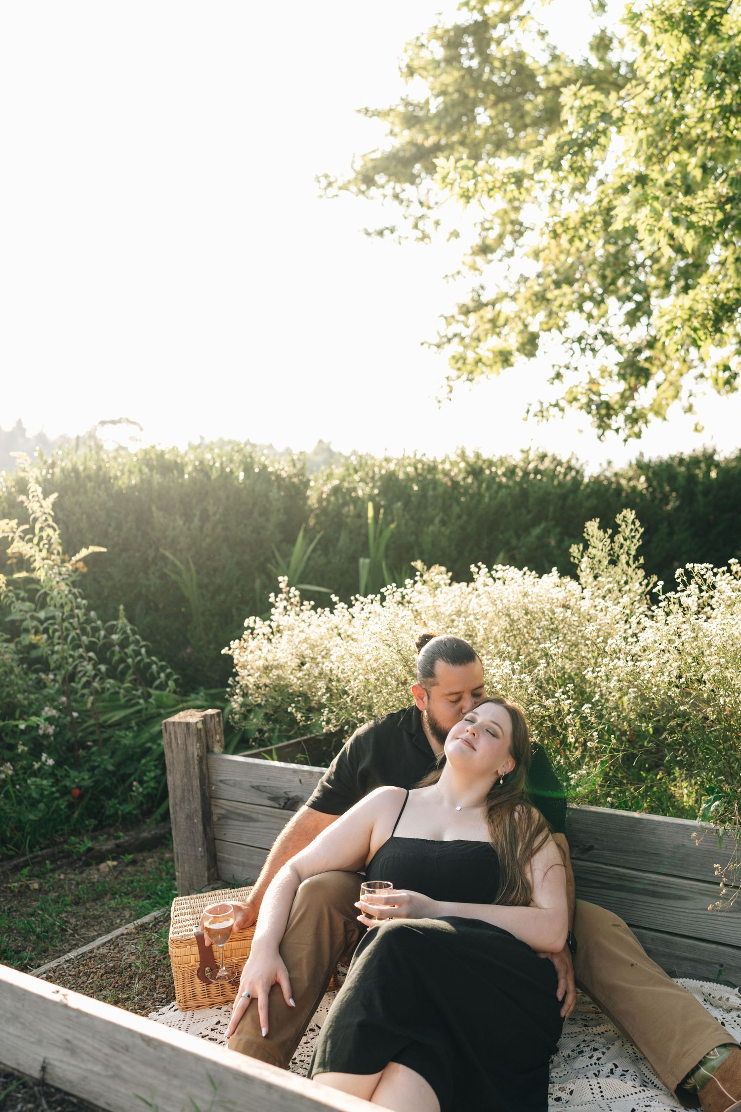 Couple relaxing on a wooden garden bench surrounded by wildflowers at sunset with a picnic basket nearby.