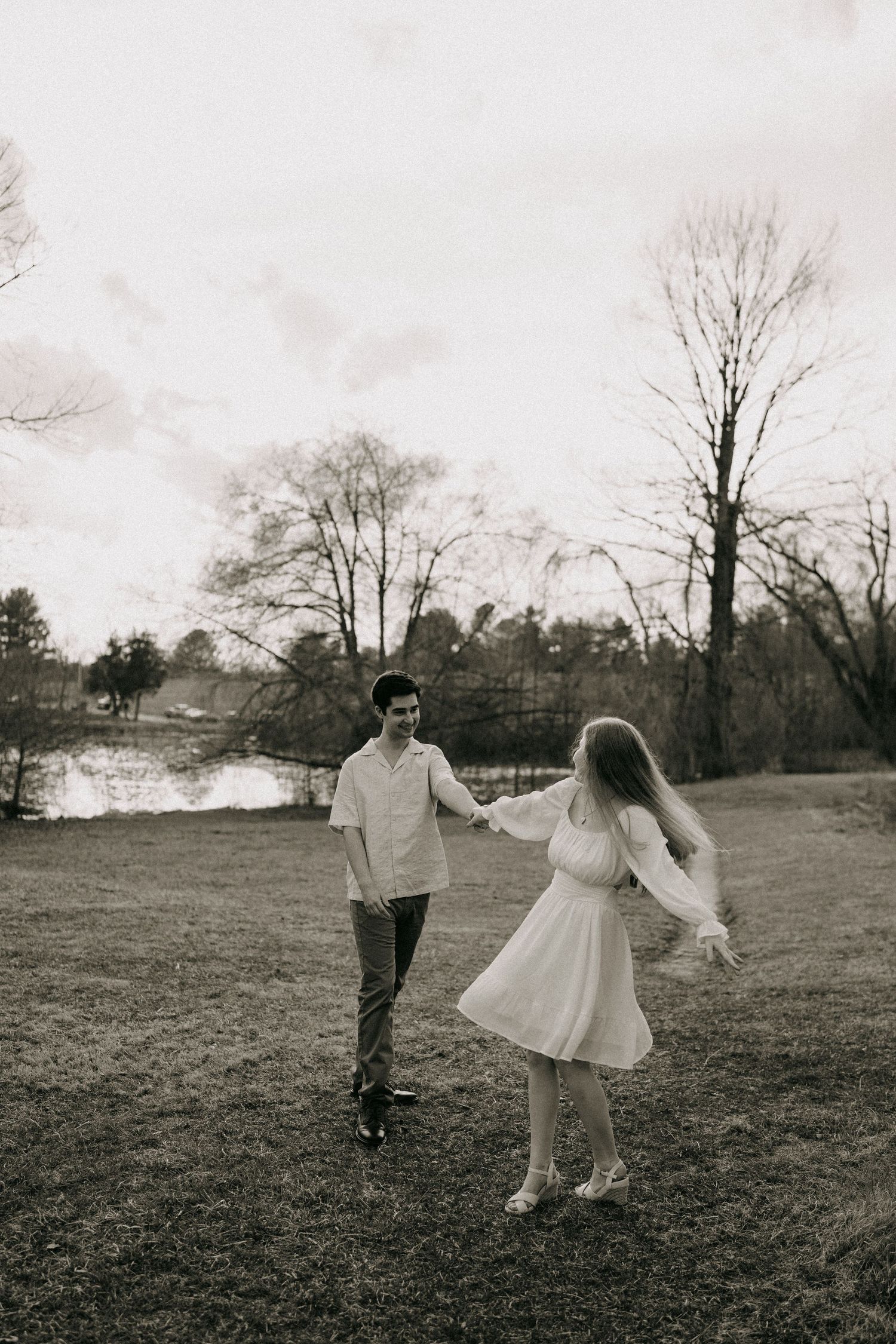 A couple holds hands and dances together in a black and white photo in a grassy park near a pond.
