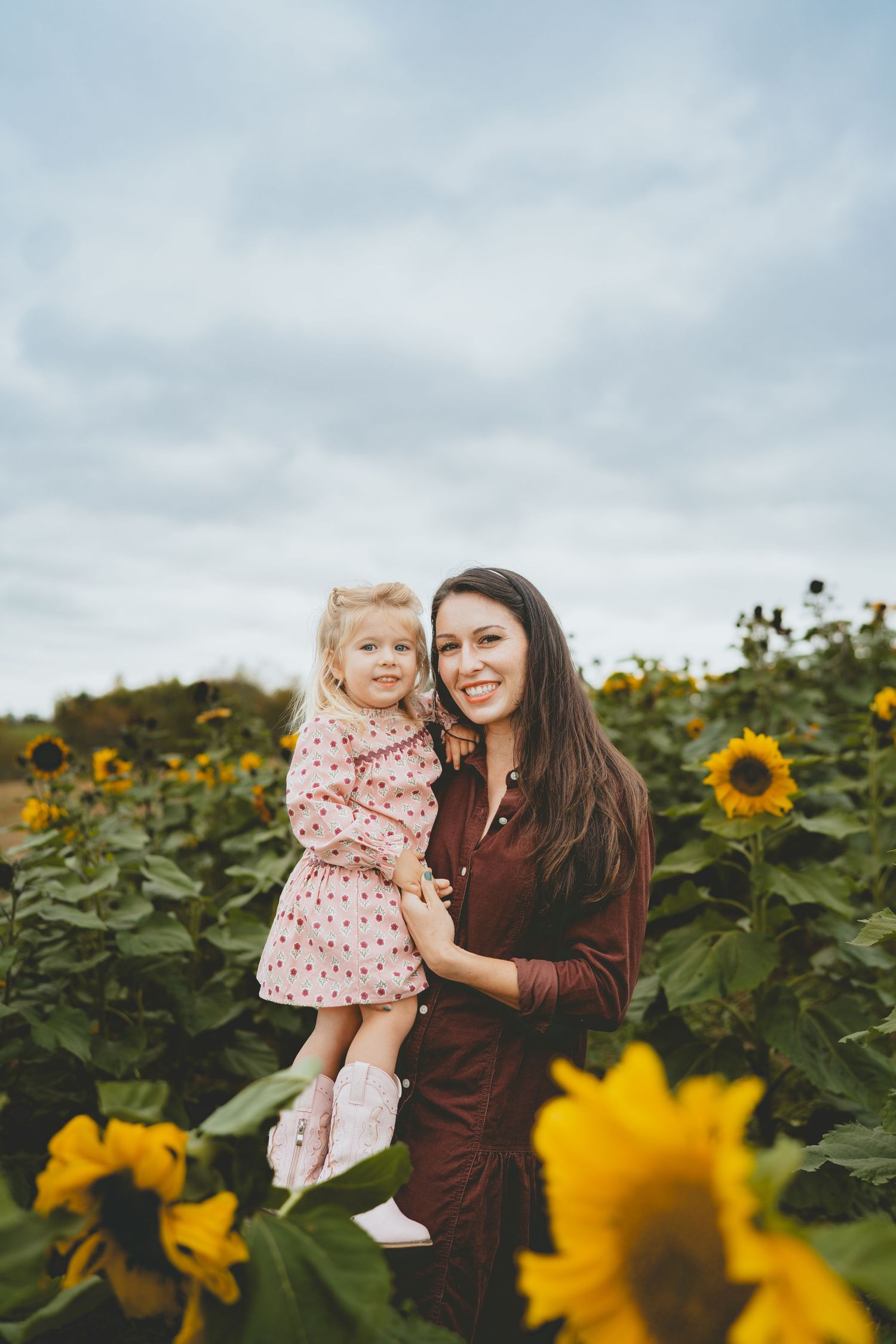 Mother holding toddler daughter in pink dress in sunflower field.