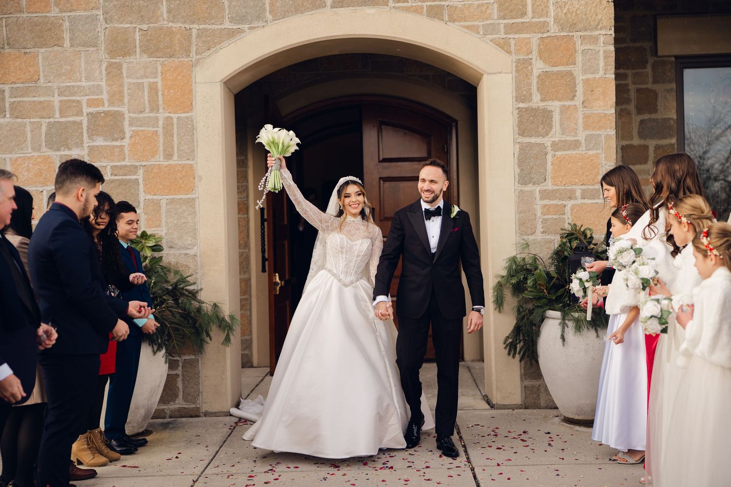 Newlyweds exit stone church as guests celebrate bride raising white bouquet in triumph.