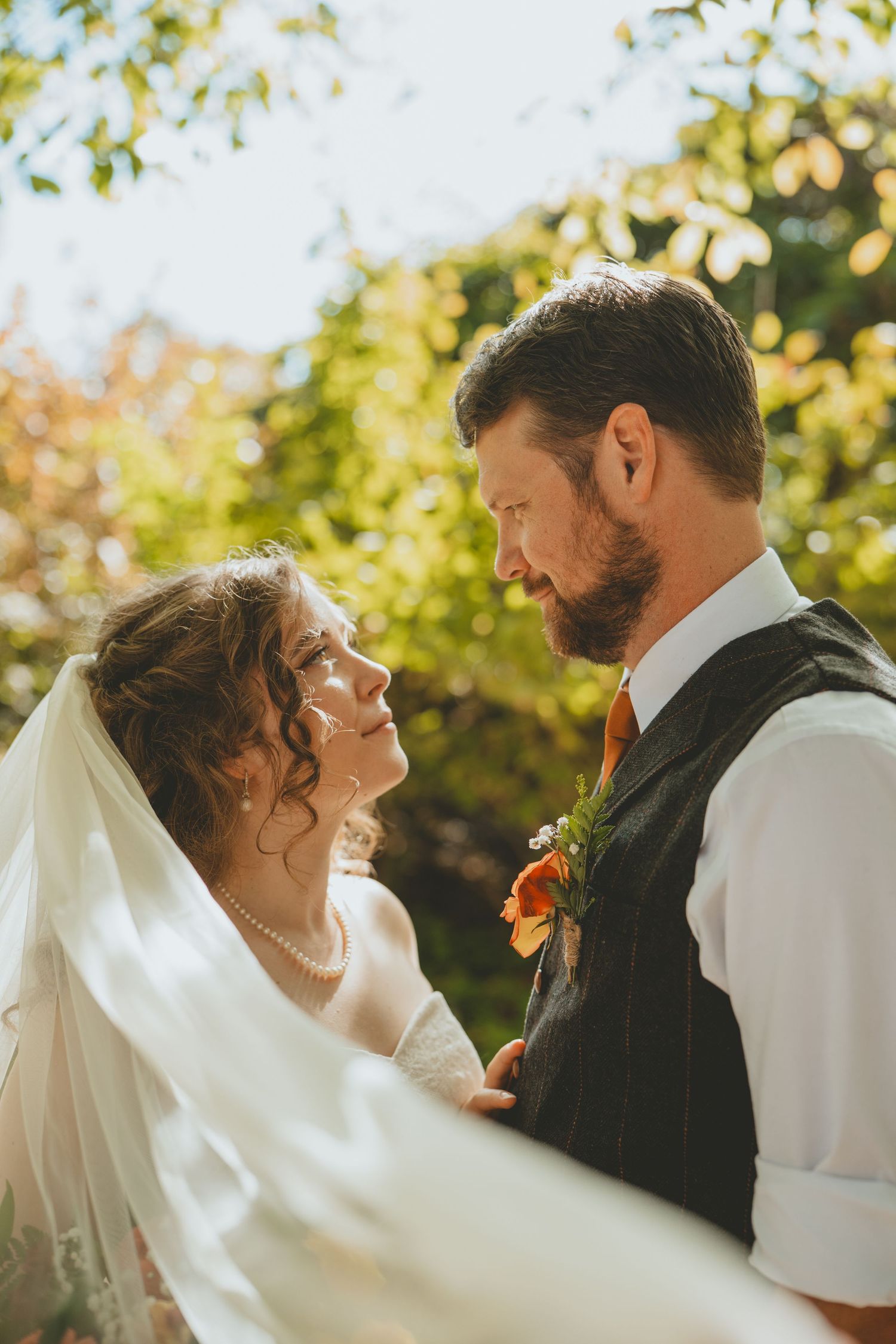 Bride and groom gazing into each other eyes under a flowing veil in warm golden sunlight outdoors.