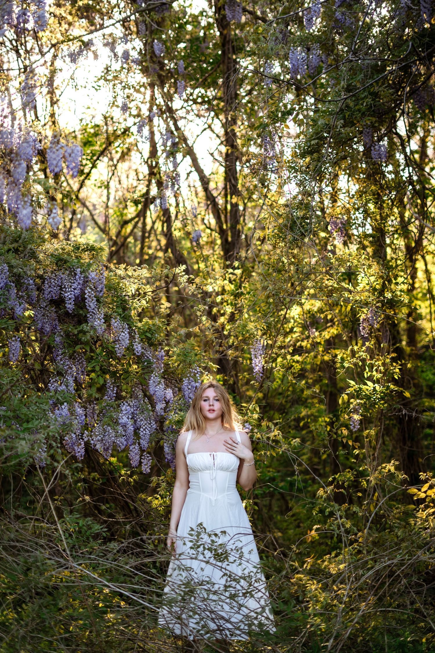 Blonde woman in white strapless gown stands surrounded by purple wisteria blooms in a sunlit forest.