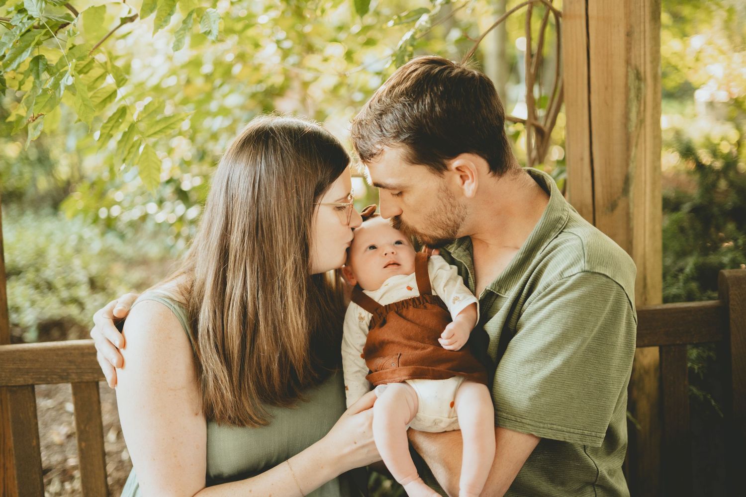 A couple kisses their newborn baby on the cheeks while standing outdoors surrounded by lush green foliage.