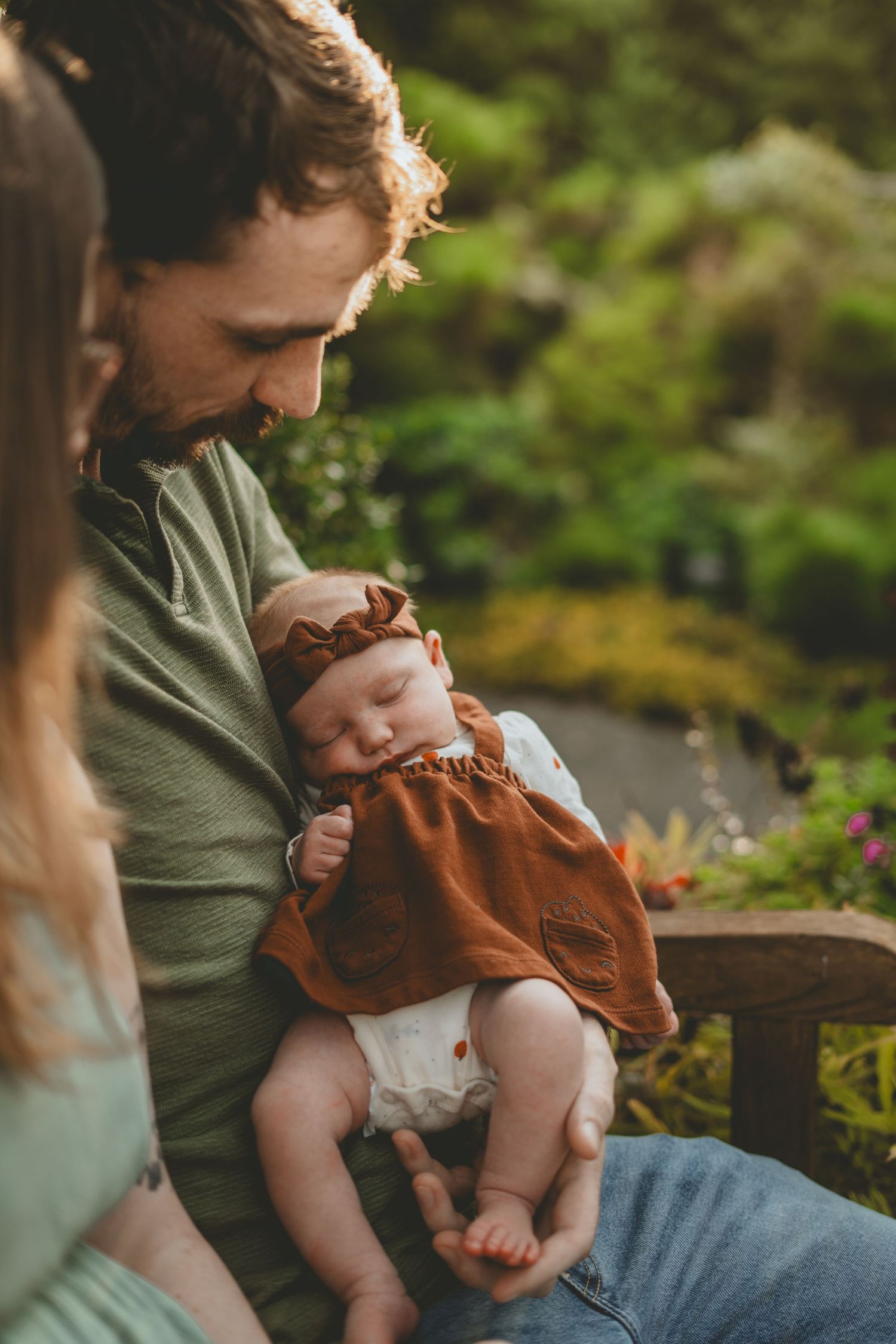 Father cradles sleeping newborn in brown dress while sitting outdoors in lush green garden.