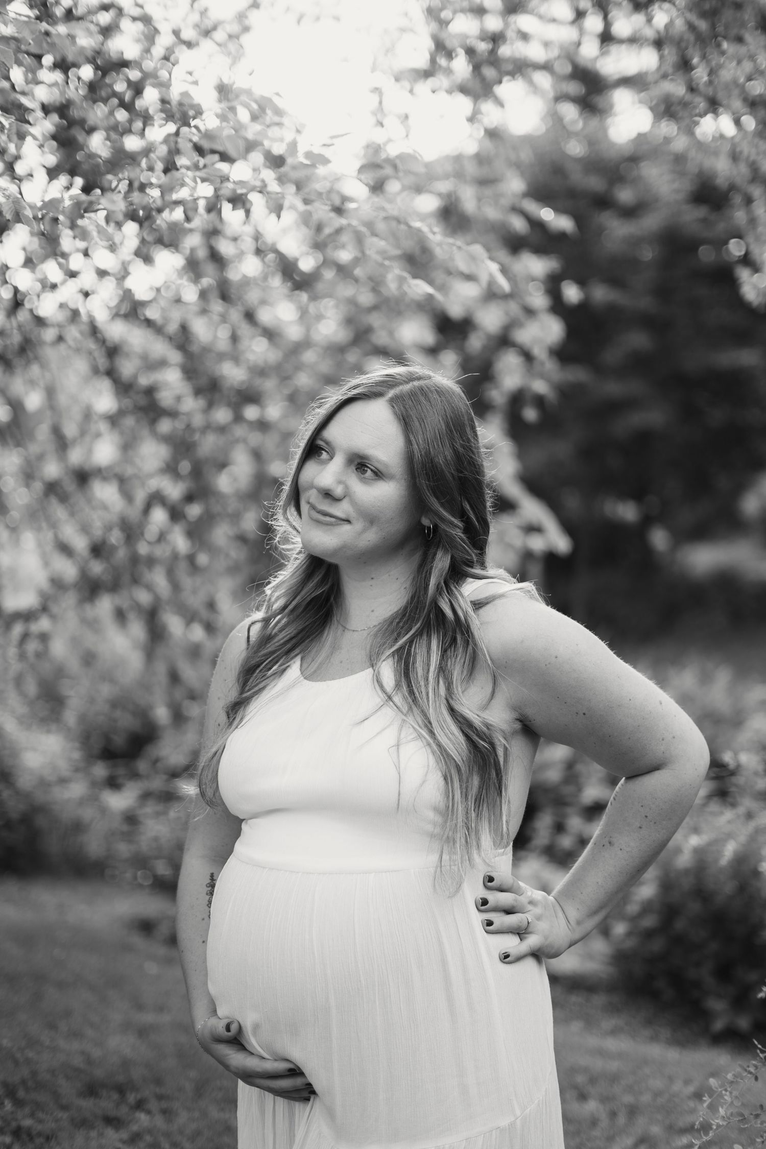 Black and white portrait of smiling pregnant woman in white dress outdoors.