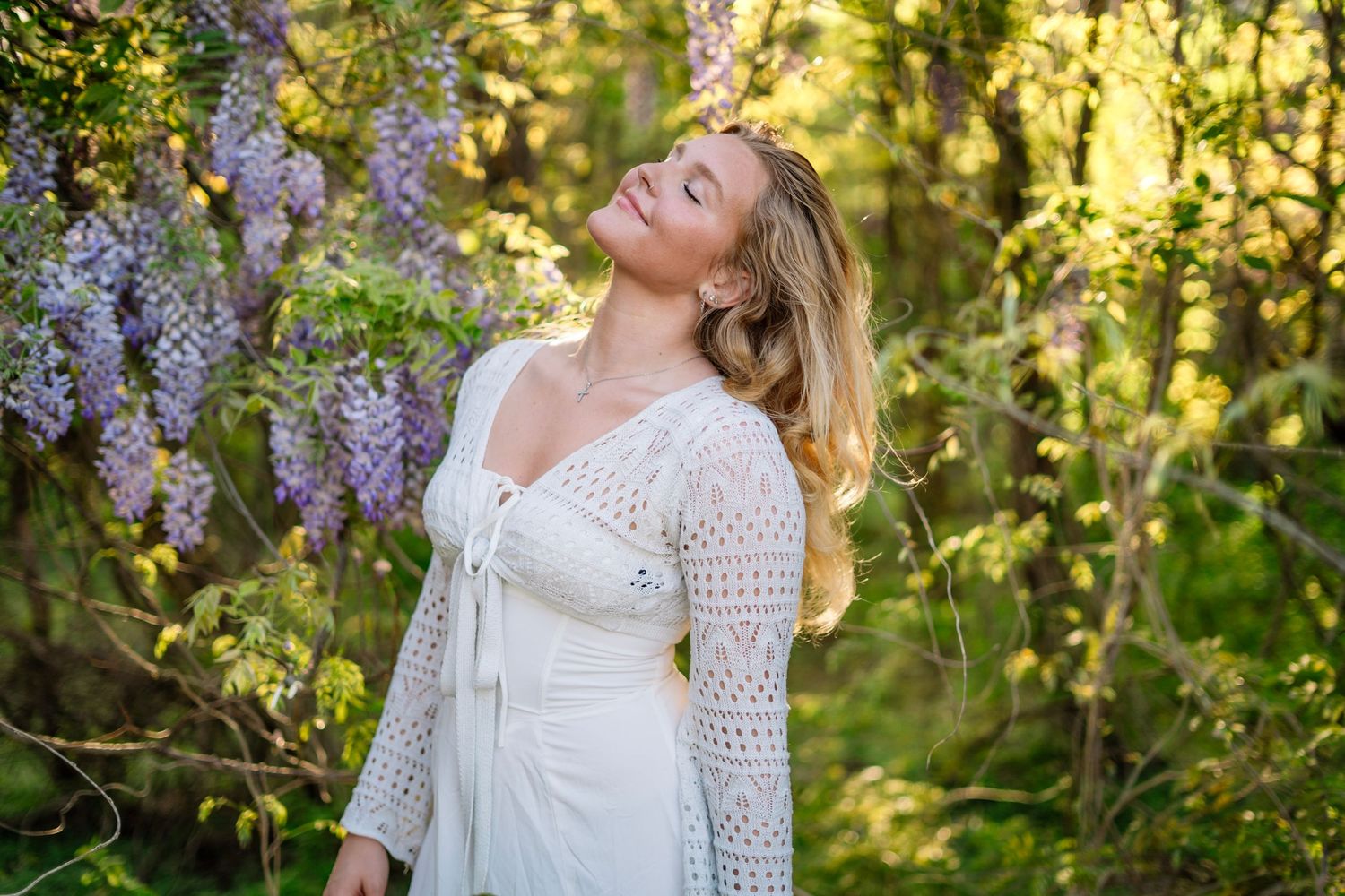 Woman in white lace dress enjoying sunlight among purple wisteria blossoms.