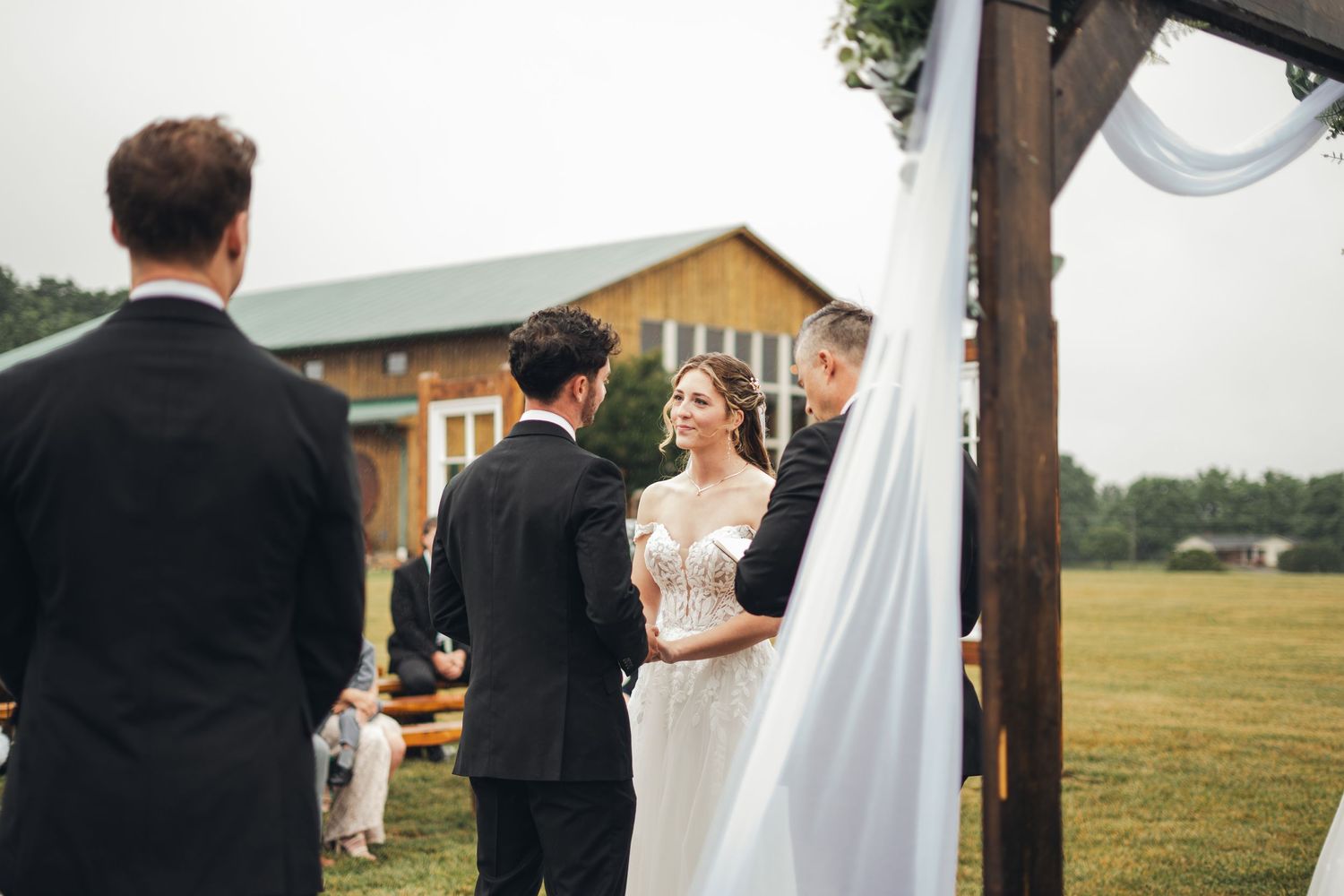 Outdoor wedding ceremony with bride in white gown and groom in black suit under wooden arch, rustic barn in background.
