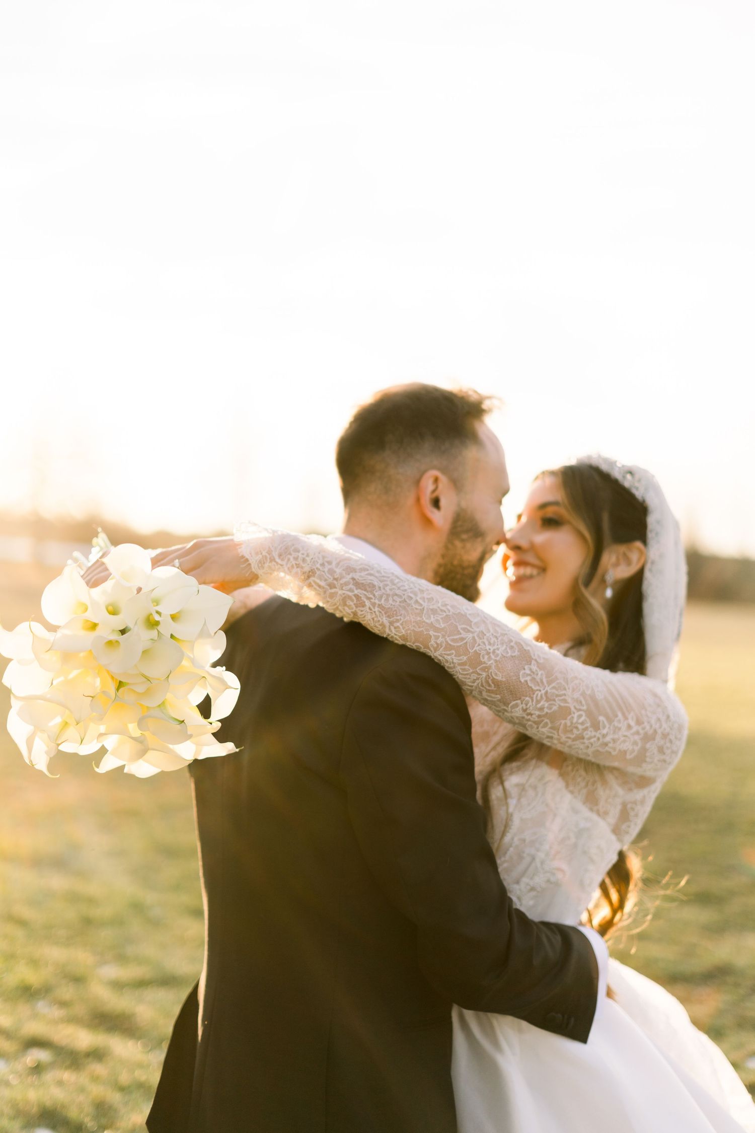 Bride in lace gown embraces groom while holding white bouquet, both smiling in a glowing sunset field.