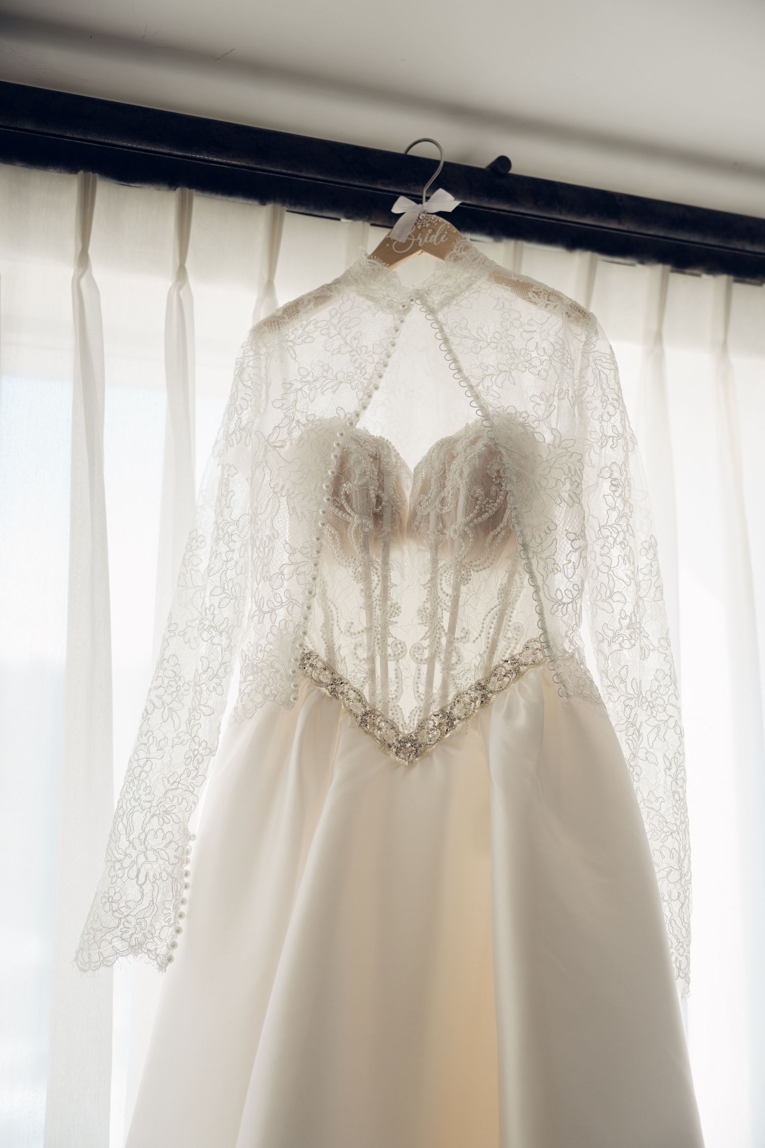 Elegant white wedding dress with lace cape and pearl buttons hanging on a wooden hanger.