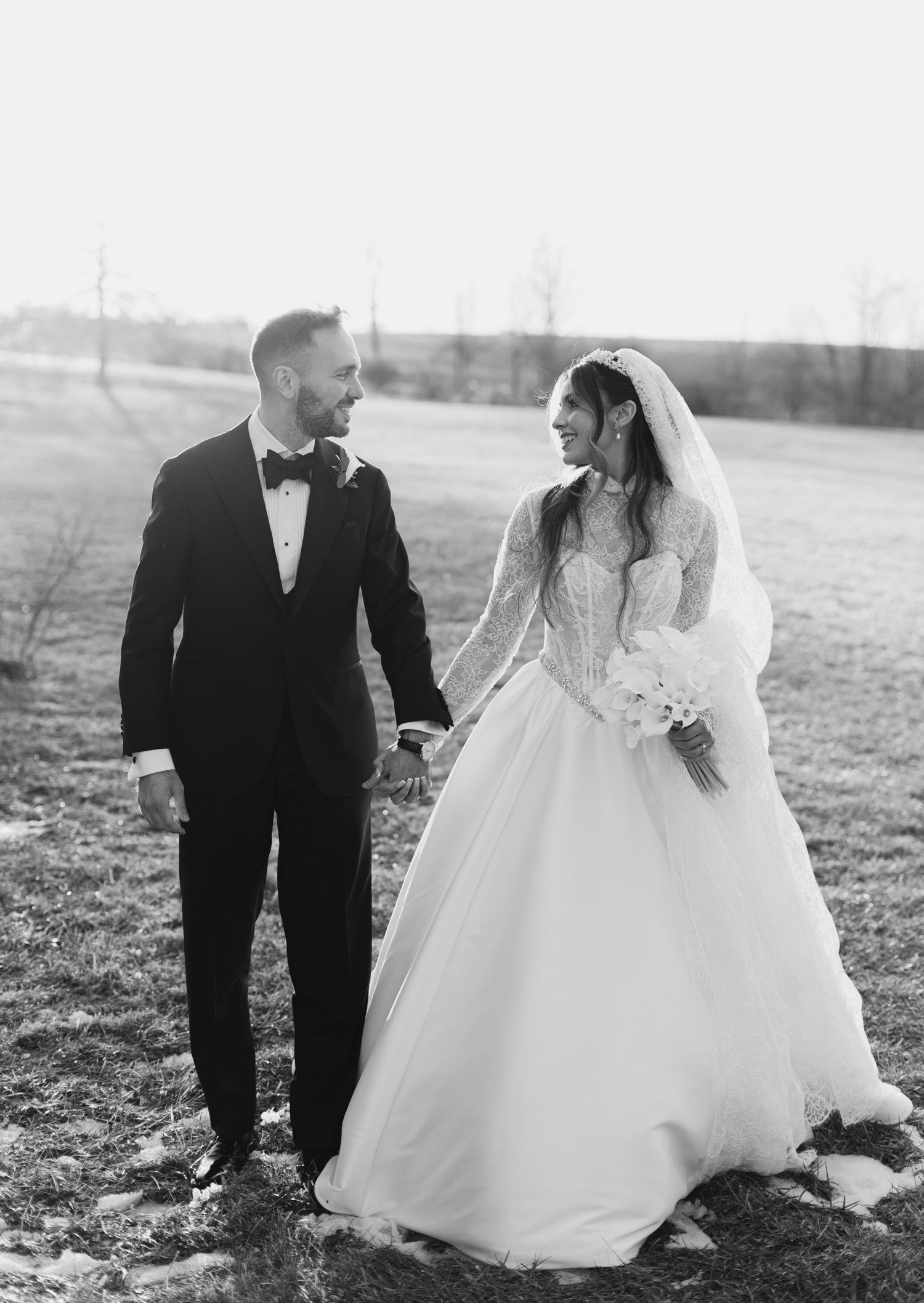Black and white photo of bride and groom holding hands and smiling while walking through a scenic outdoor field.
