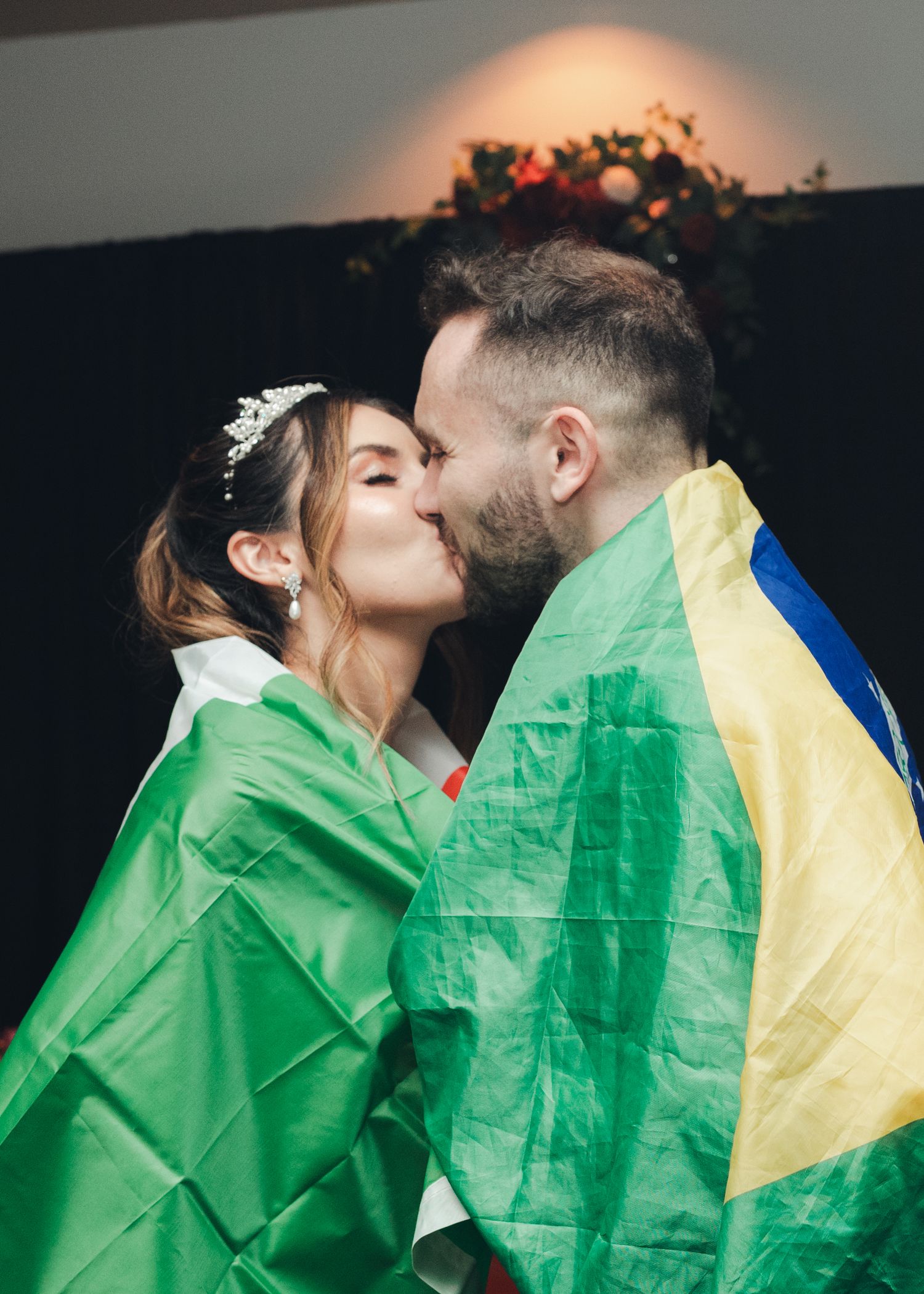 Bride and groom share a kiss while wrapped in colorful national flags at wedding reception.