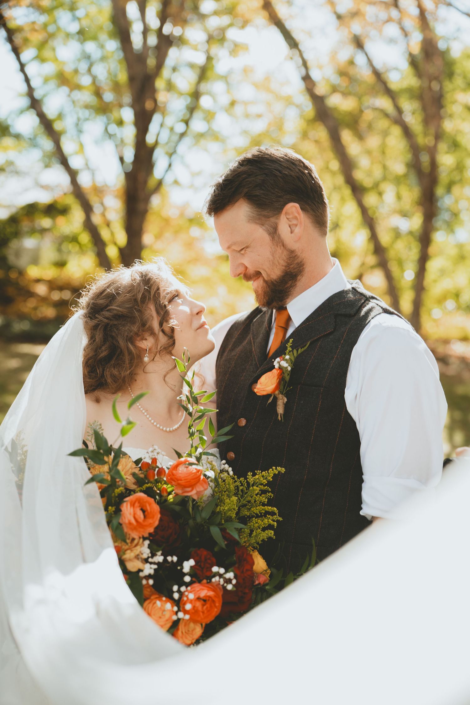 Bride and groom gazing at each other smiling, with an orange rose boutonniere and fall floral bouquet outdoors.