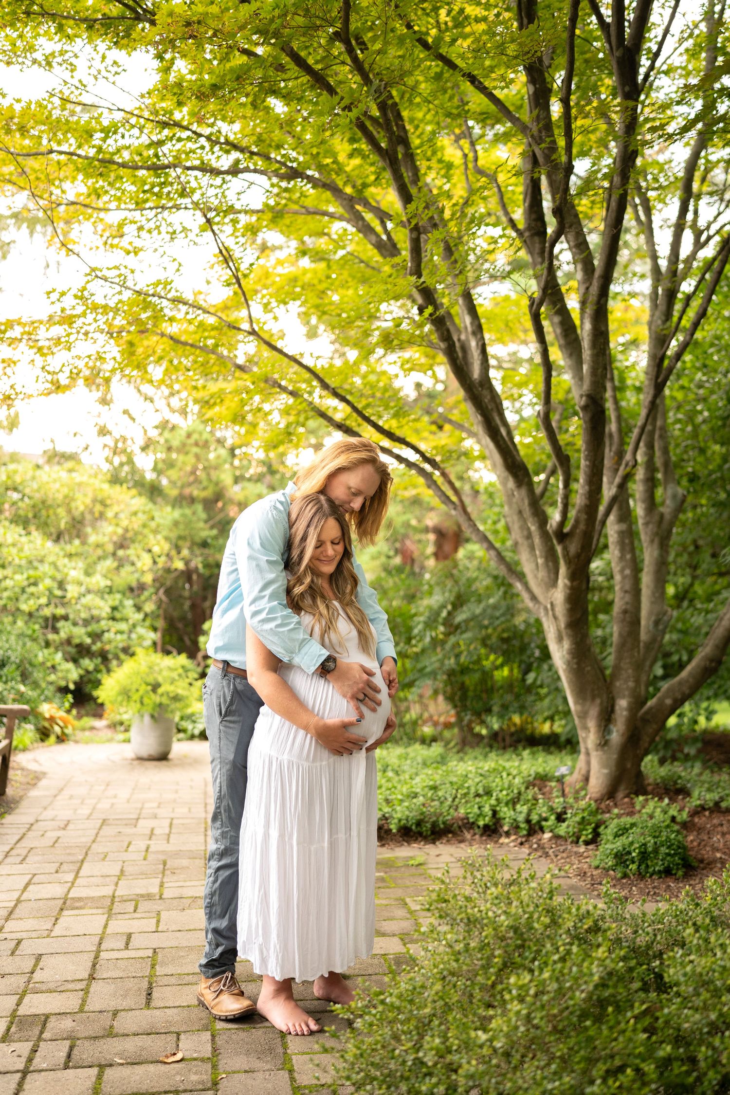 Expectant couple embracing on brick path under sunlit green trees in garden.