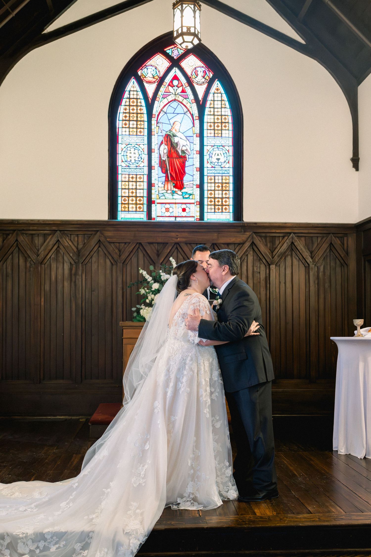 Bride and groom share first kiss at altar beneath vibrant stained glass window in historic chapel.